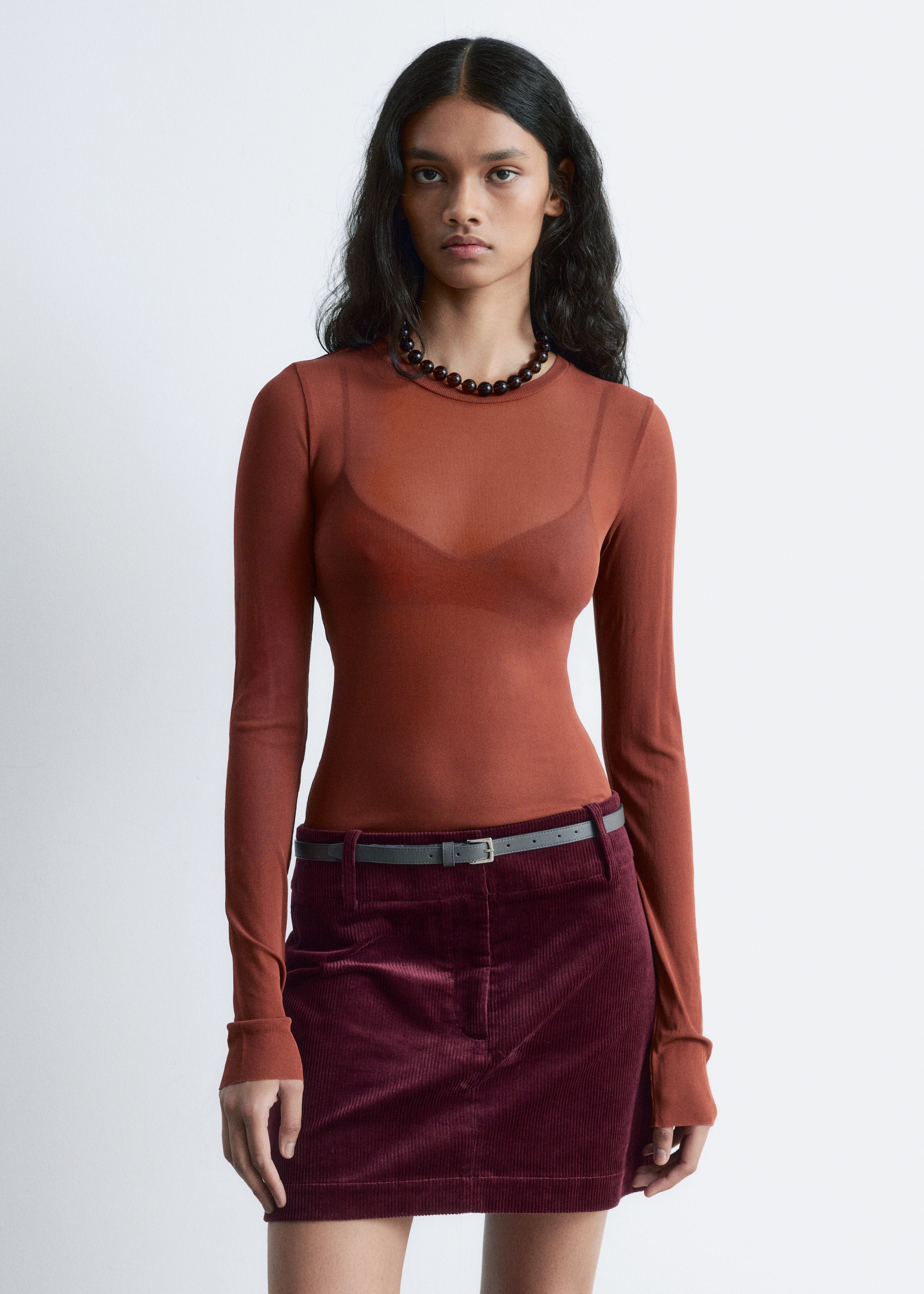 View larger image: A form-fitting, sheer, rust-colored long-sleeved top is being worn, with a dark bra visible underneath. A fitted, dark red corduroy mini skirt is also being worn, accented by a thin grey belt.