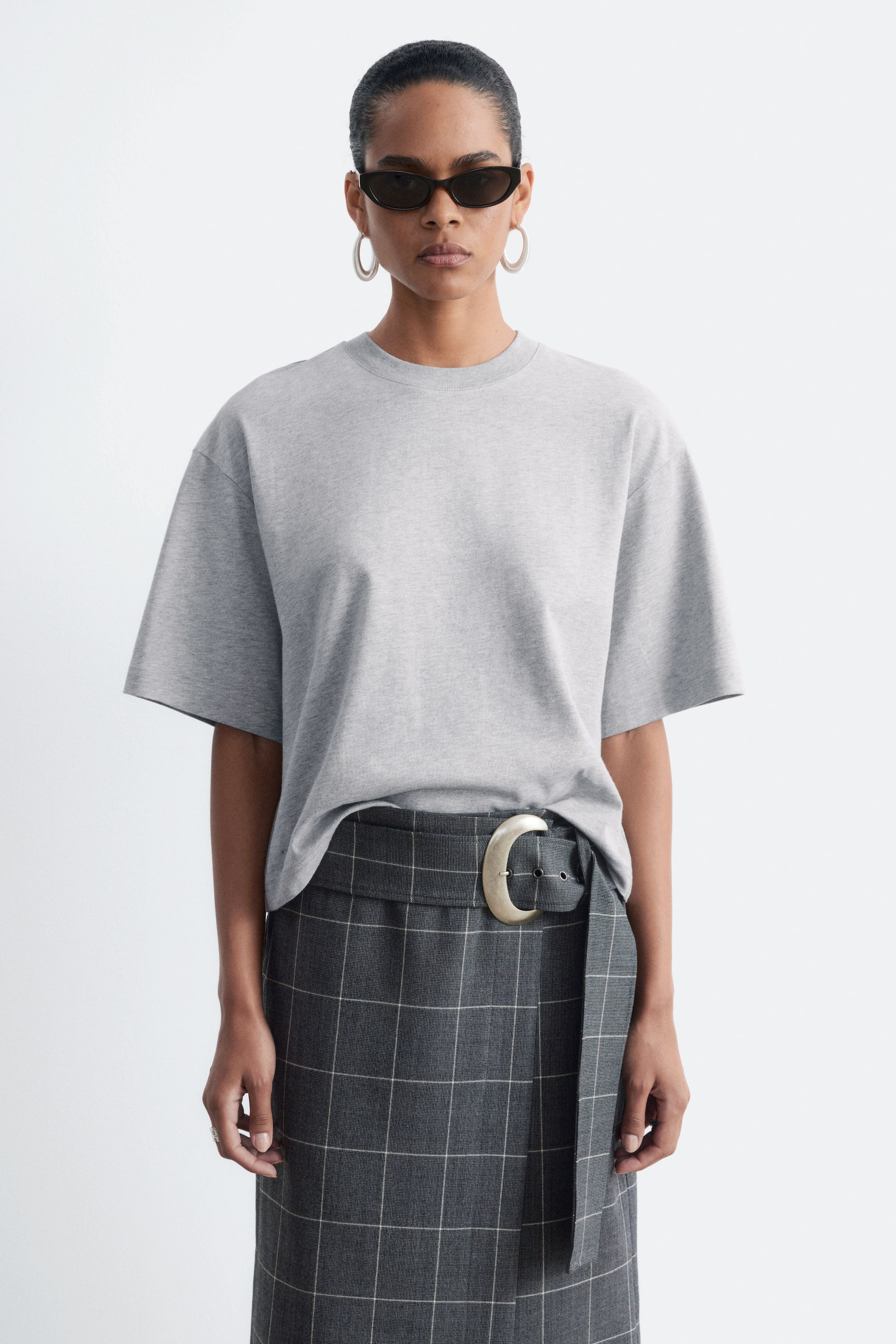 Agrandir l'image: Un t-shirt gris clair à la coupe ample et carrée, avec un col rond et de larges manches courtes, est porté par le mannequin. Le t-shirt est porté avec l’ourlet avant rentré dans une jupe grise à carreaux taille haute, ornée d’une grande boucle de ceinture métallique.