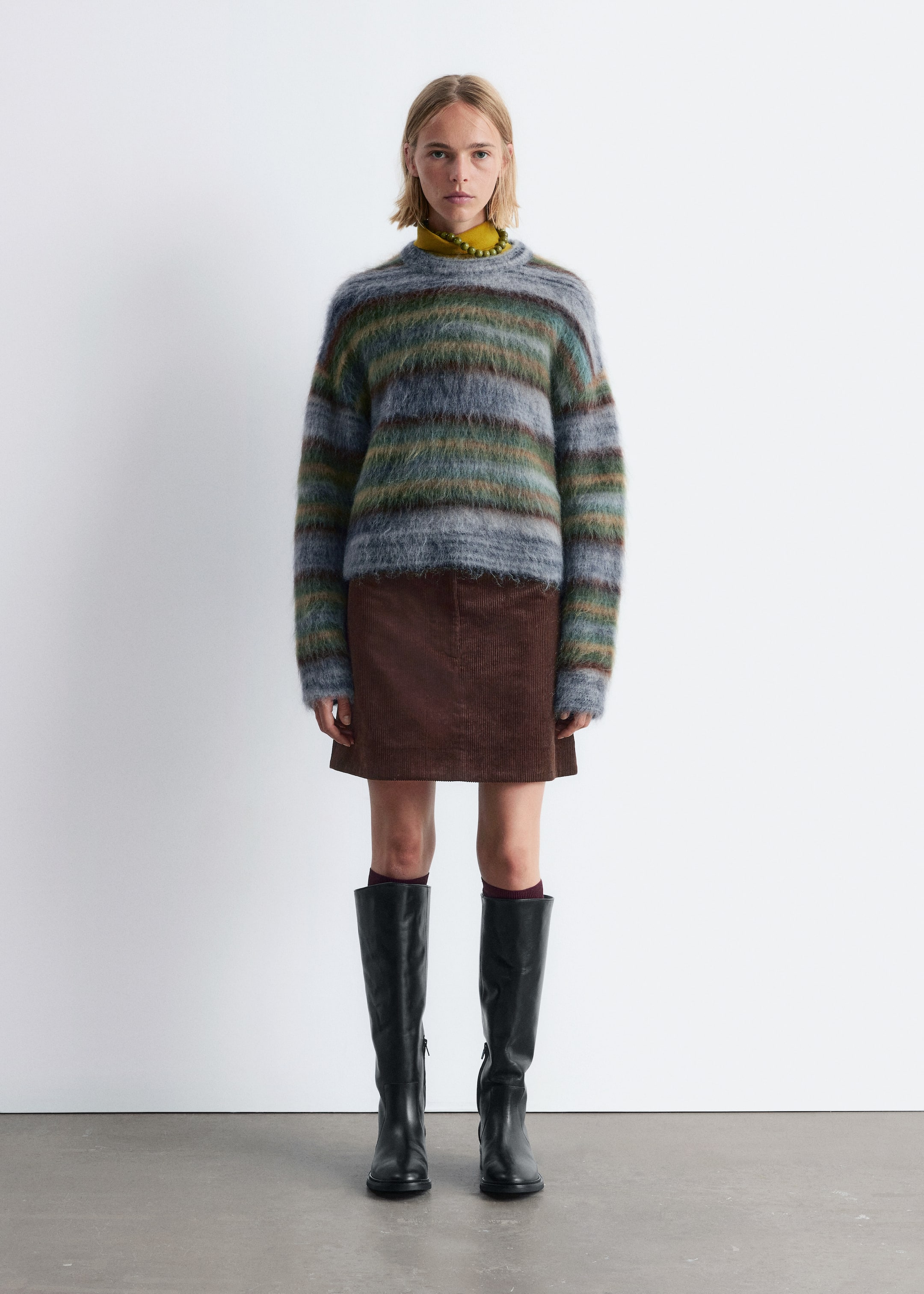 Ingrandisci l'immagine: Un maglione a girocollo a righe orizzontali e sfumature di grigio, blu, verde e marrone viene indossato sopra un dolcevita giallo. È abbinato a una minigonna a trapezio in velluto a coste marrone e stivali neri al ginocchio rivolti in avanti, che rivelano calzini bordeaux.