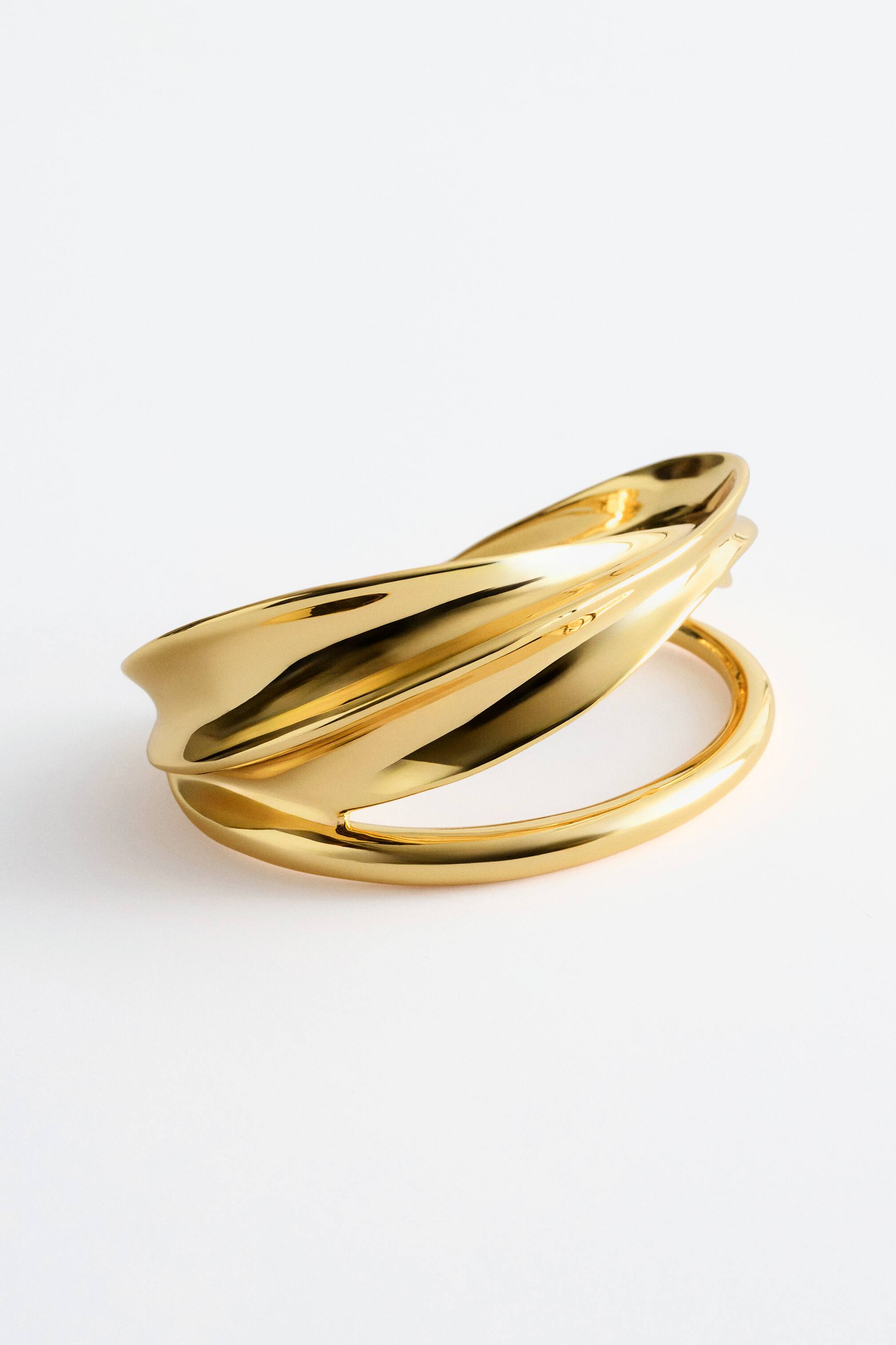 Visa större bild: Ett manschettarmband, tillverkat av polerad mässing, visar upp sin skulpturala, tvålagersdesign, presenterad framifrån. Det översta lagret bildar en vågig, flöjtformad form, medan det nedre lagret är ett öppet, rundat band.