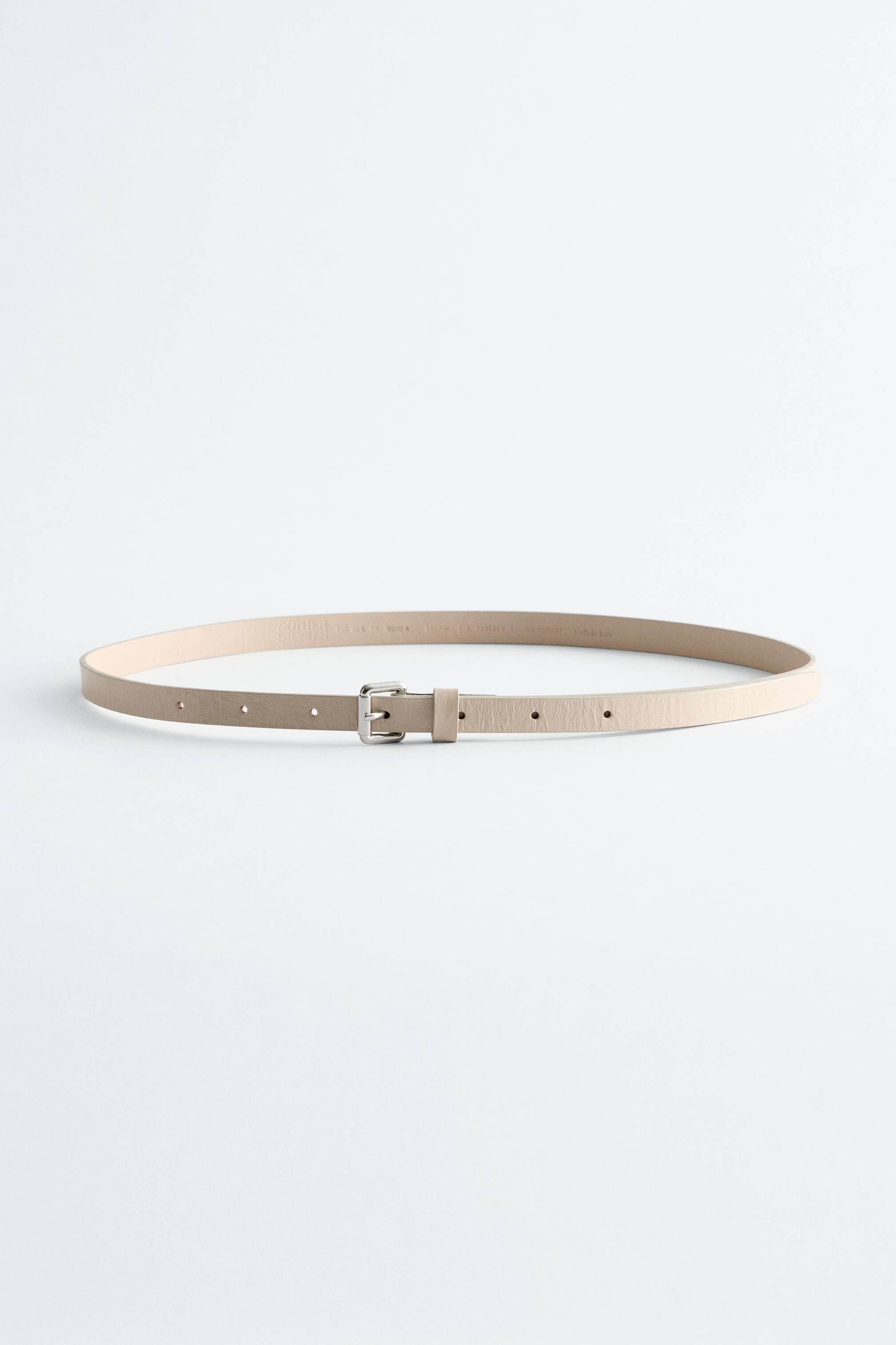 Visualizza immagine più grande: Una sottile cintura in pelle beige chiaro con texture si curva in una forma ovale, presentando la sua fibbia quadrata argentata nella parte anteriore. Piccoli fori rotondi sono visibili lungo la lunghezza della cintura.