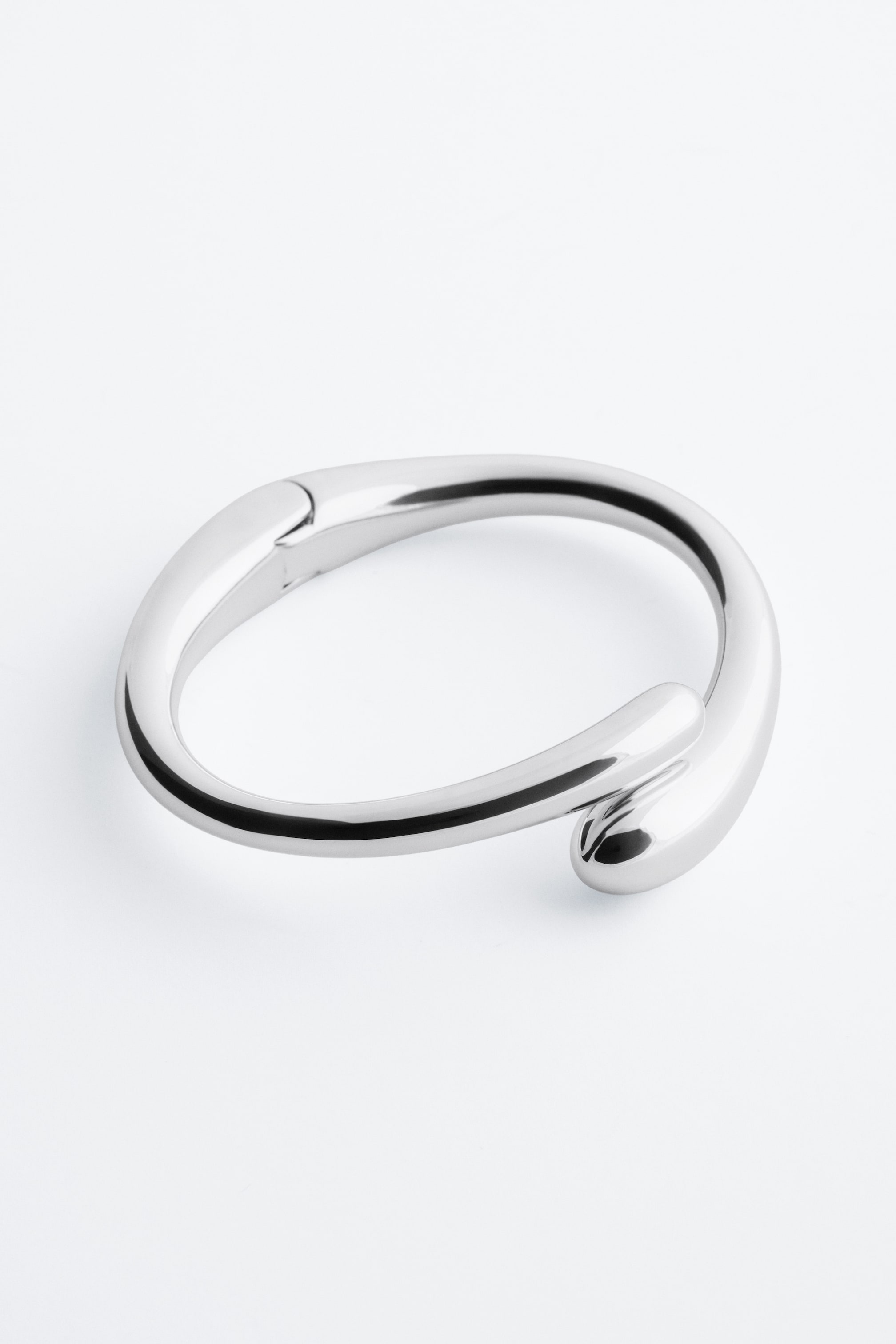 Visa större bild: Ett skulpturalt manschettarmband i polerad zink visar upp sin form med vidgande ändar. Det har ett gångjärnsstängning och den ena änden böjer sig över den andra.