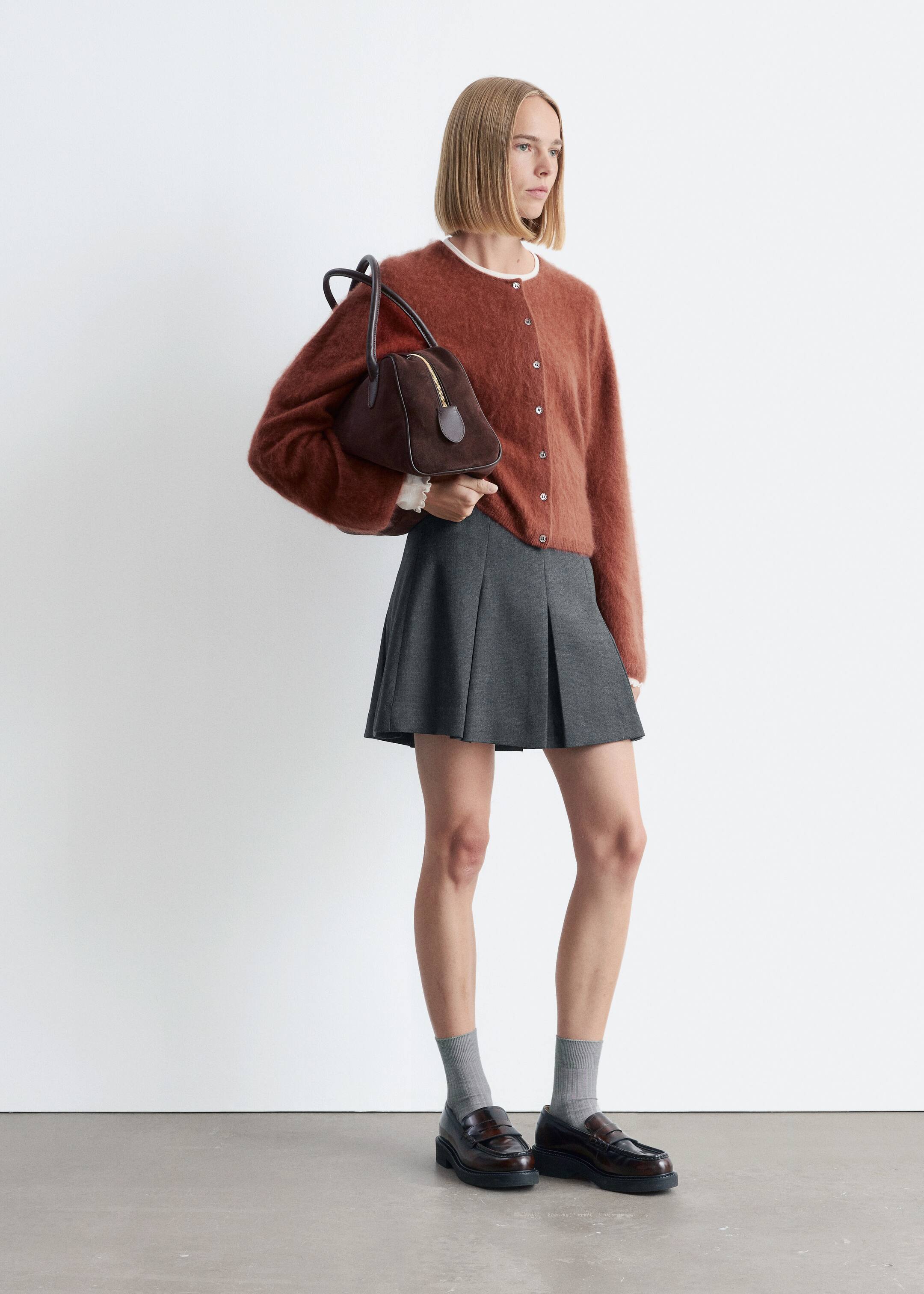 Größeres Bild anzeigen: Eine rostfarbene gestrickte Kaschmir-Strickjacke mit flauschiger Textur und Knopfleiste wird vom Model über einem weißen Oberteil getragen. Das Model hält eine dunkelbraune Handtasche.