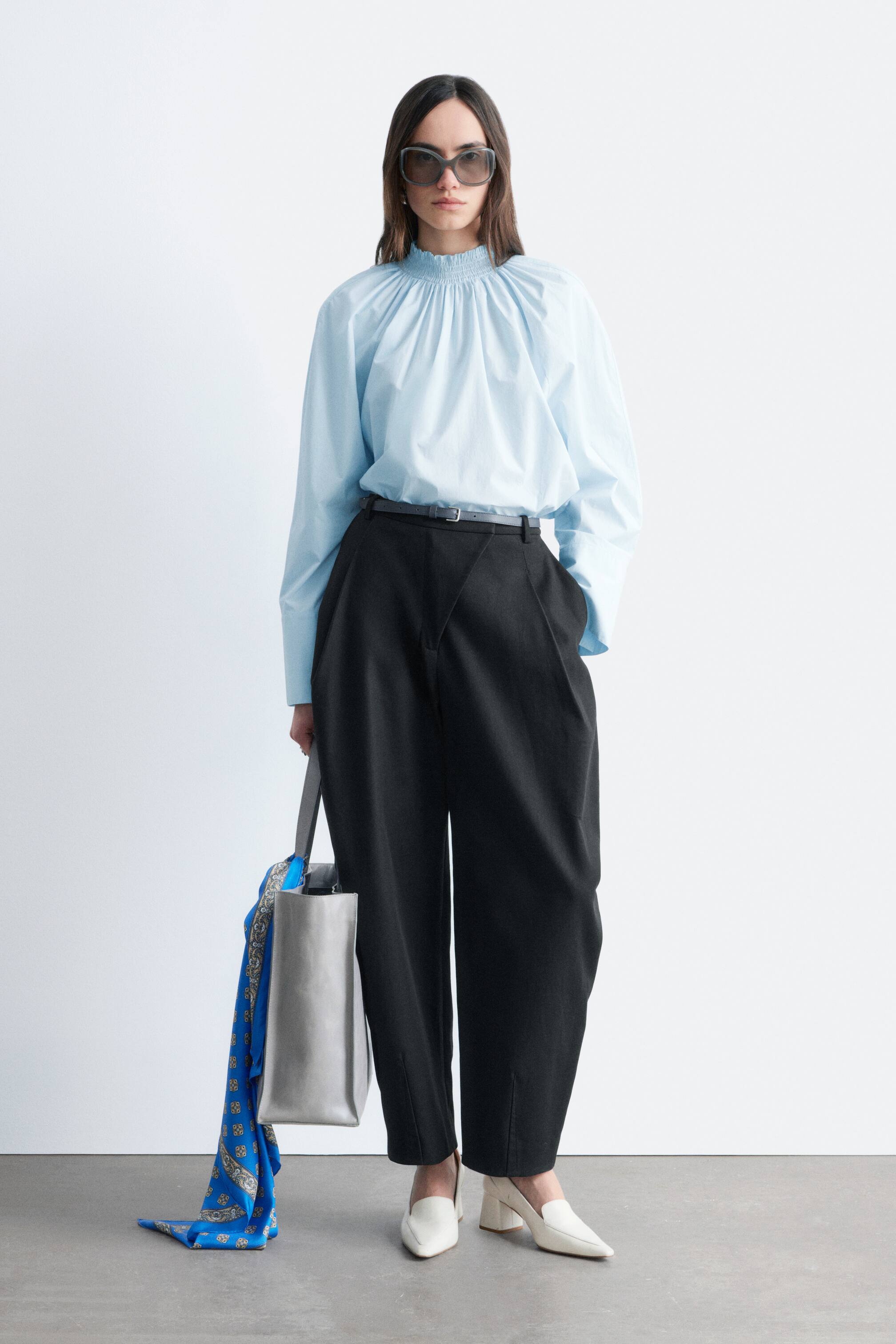 Visualizza immagine più grande: La modella indossa una camicetta in popeline di cotone azzurro chiaro, caratterizzata da un colletto a imbuto arricciato e pieghe nette. La camicetta presenta polsini con bottoni ed è abbinata a pantaloni scuri con cintura.