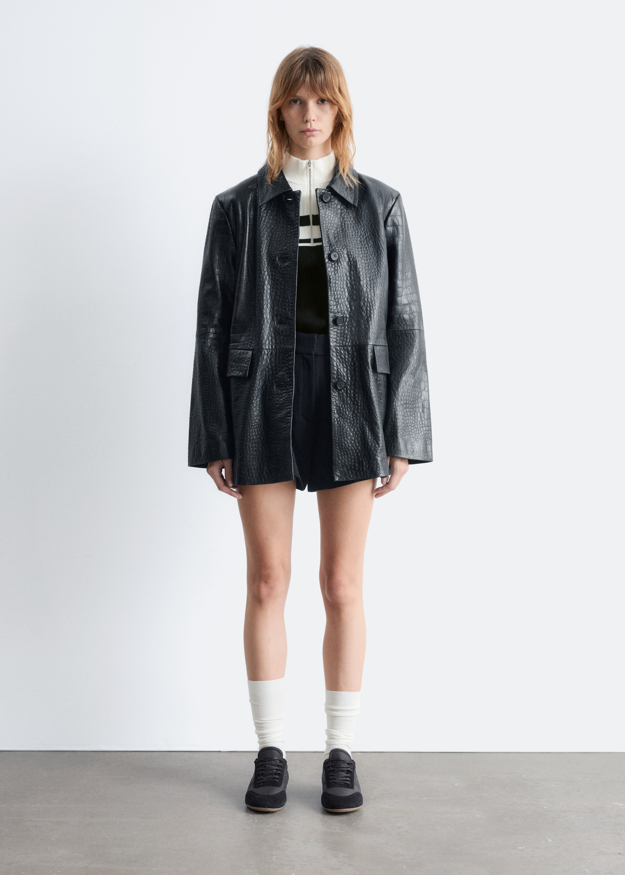 Agrandir l'image: Une veste embossée en crocodile noir est présentée face avant, avec un col pointu, une fermeture à boutons à l'avant et deux poches à rabat. Elle est portée par-dessus un pull à col montant rayé.