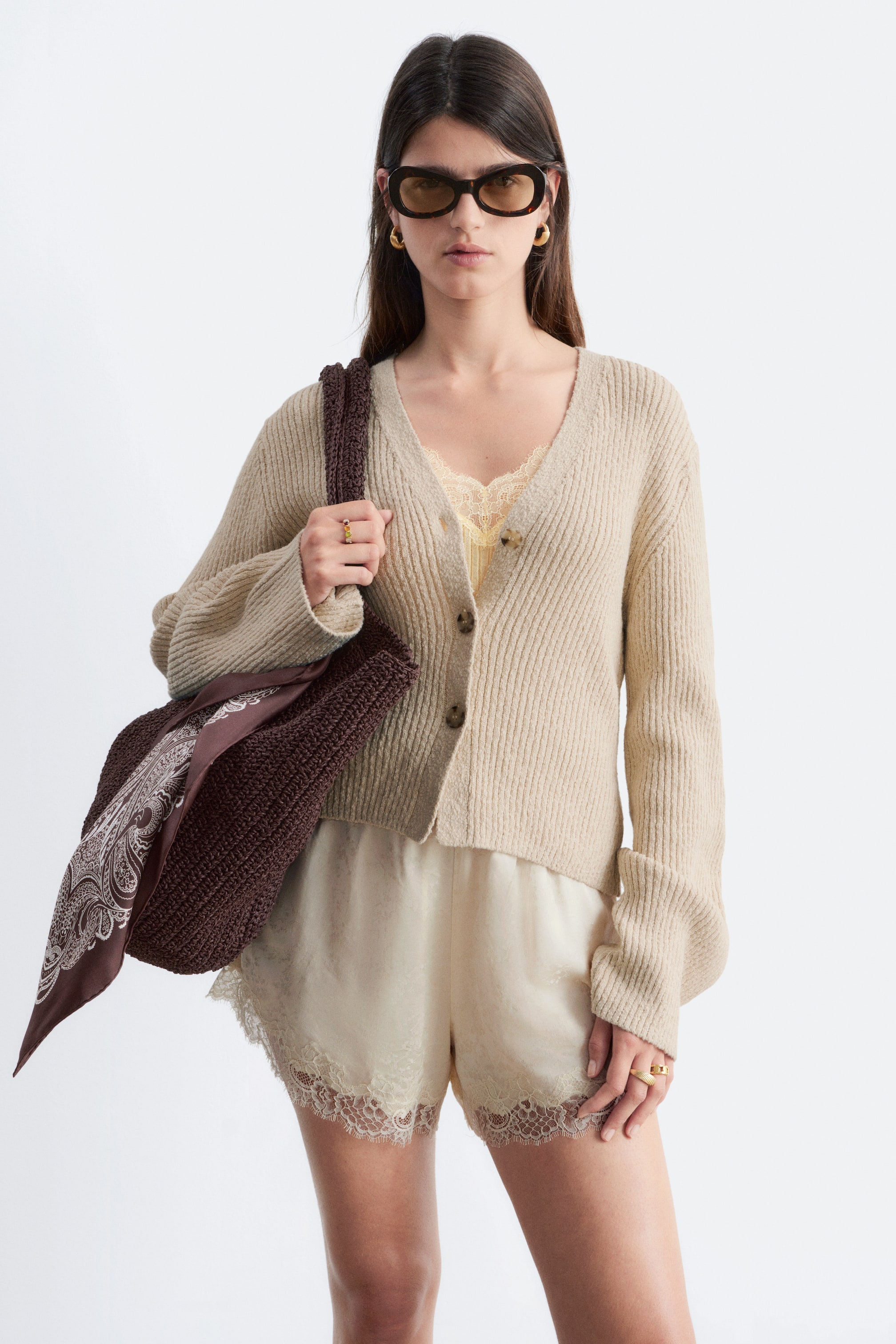 Agrandir l'image: Un cardigan en maille côtelée beige est présenté de face, mettant en avant un décolleté en V, trois boutons marron et des manches longues. Il est porté ouvert sur un caraco jaune bordé de dentelle et est assorti à un short crème, qui présente des ourlets festonnés en dentelle.