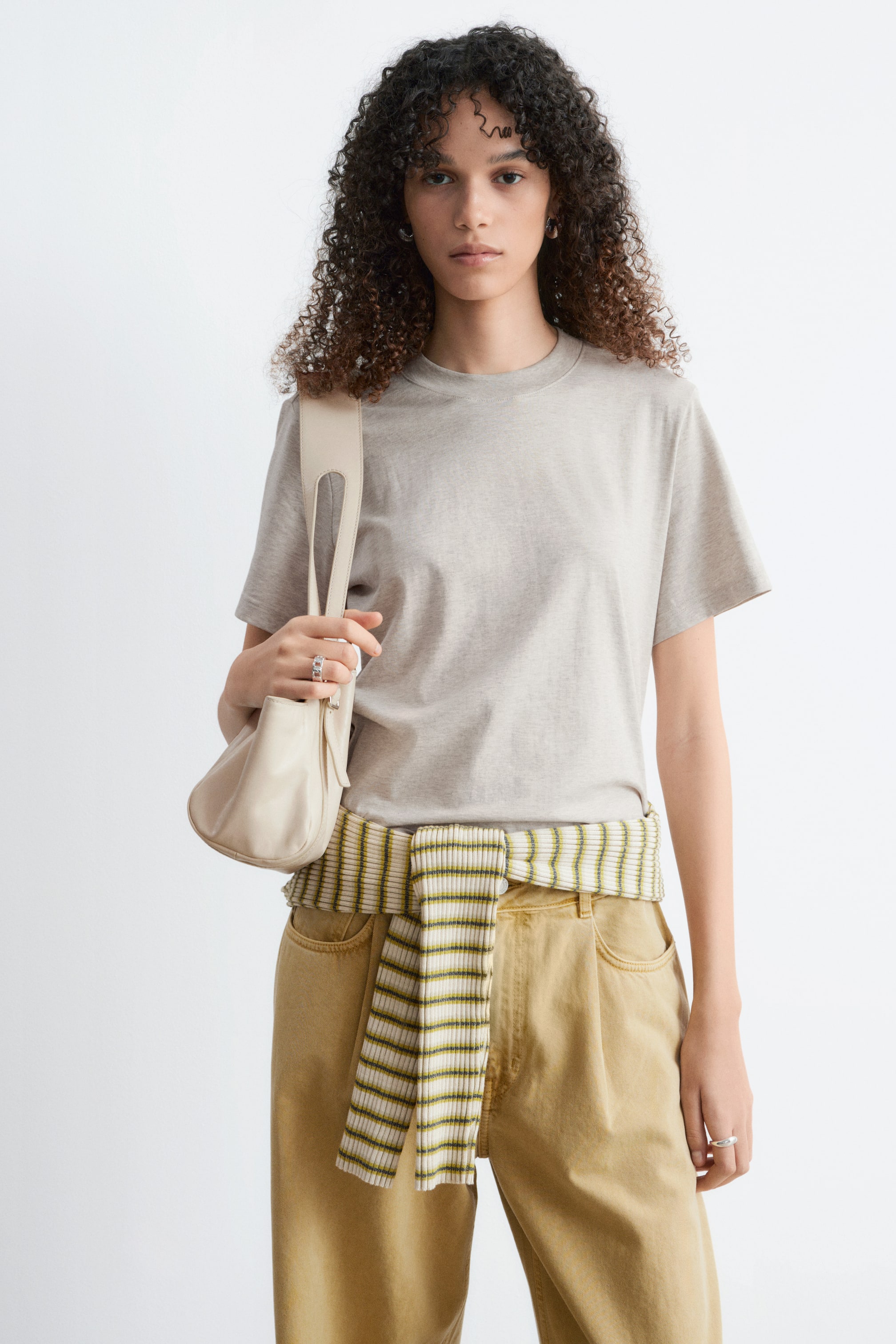 Ver imagem maior: A modelo está a usar uma t-shirt cinzenta clara de manga curta e decote redondo com uma silhueta descontraída. Uma peça de roupa às riscas verdes e brancas está amarrada à cintura da modelo, sobrepondo-se à t-shirt, que é combinada com calças bege.