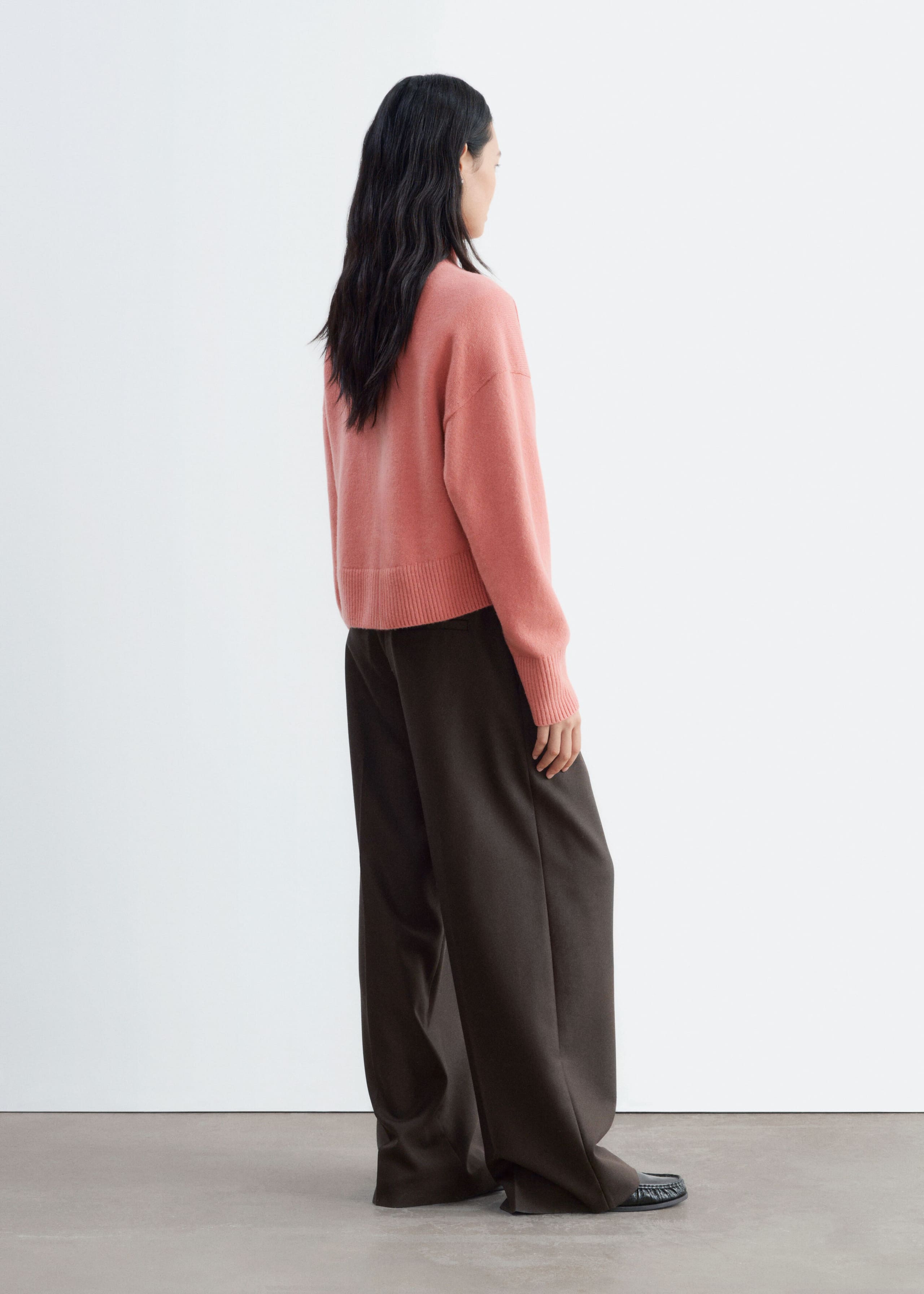 The model is wearing a pink mock-neck jumper, shown from a three-quarter back view to highlight its relaxed silhouette, dropped shoulder seams, and ribbed cuffs and hem. The jumper is paired with dark brown wide-leg trousers.