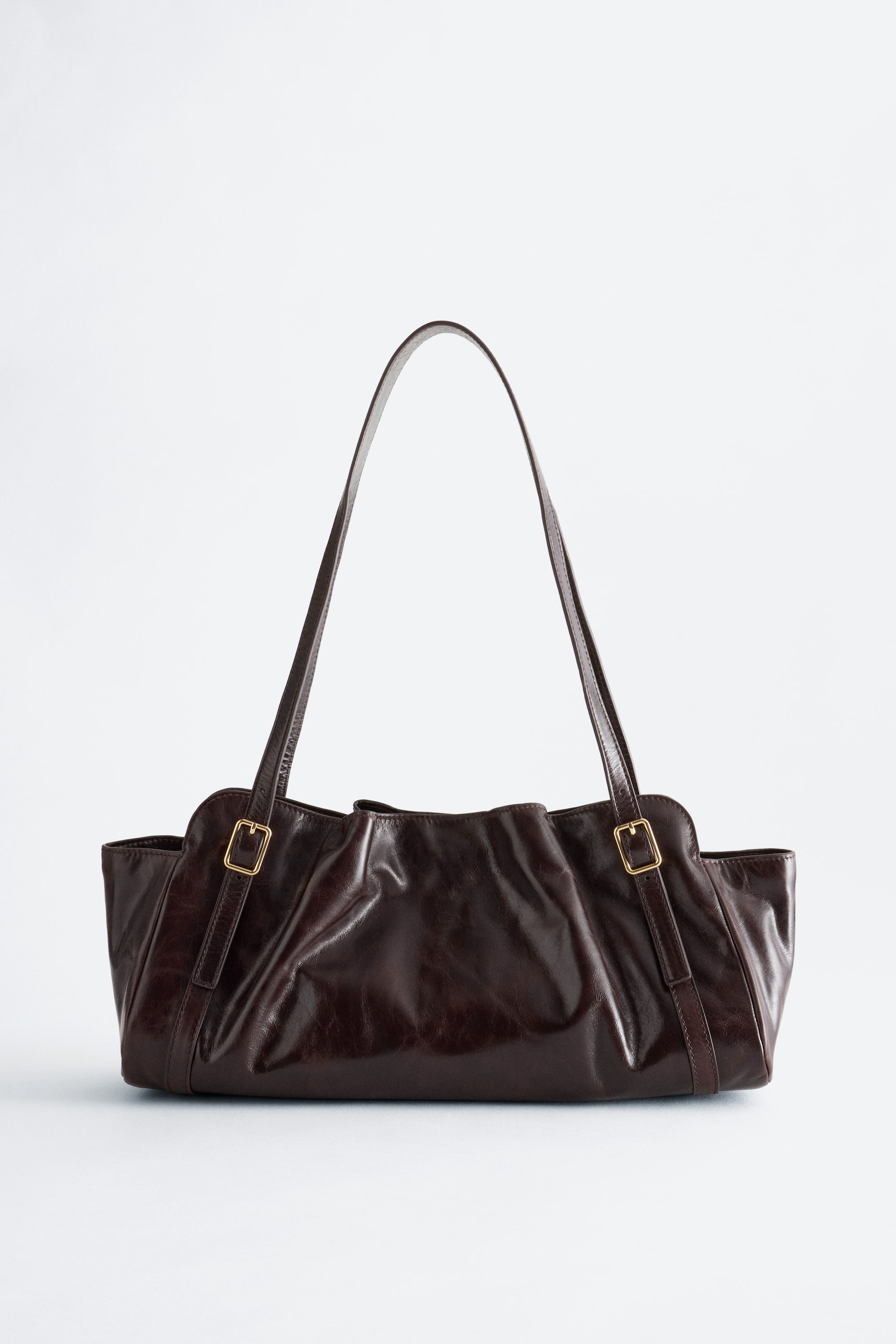 Agrandir l'image: Un sac à bandoulière en cuir brun foncé est présenté debout, de face, mettant en valeur sa silhouette allongée et discrète, avec un corps souplement plissé et légèrement affaissé. Le sac est doté d’une seule longue bandoulière fixée à l’avant par deux bandes verticales en cuir et de petites boucles rectangulaires de couleur dorée.