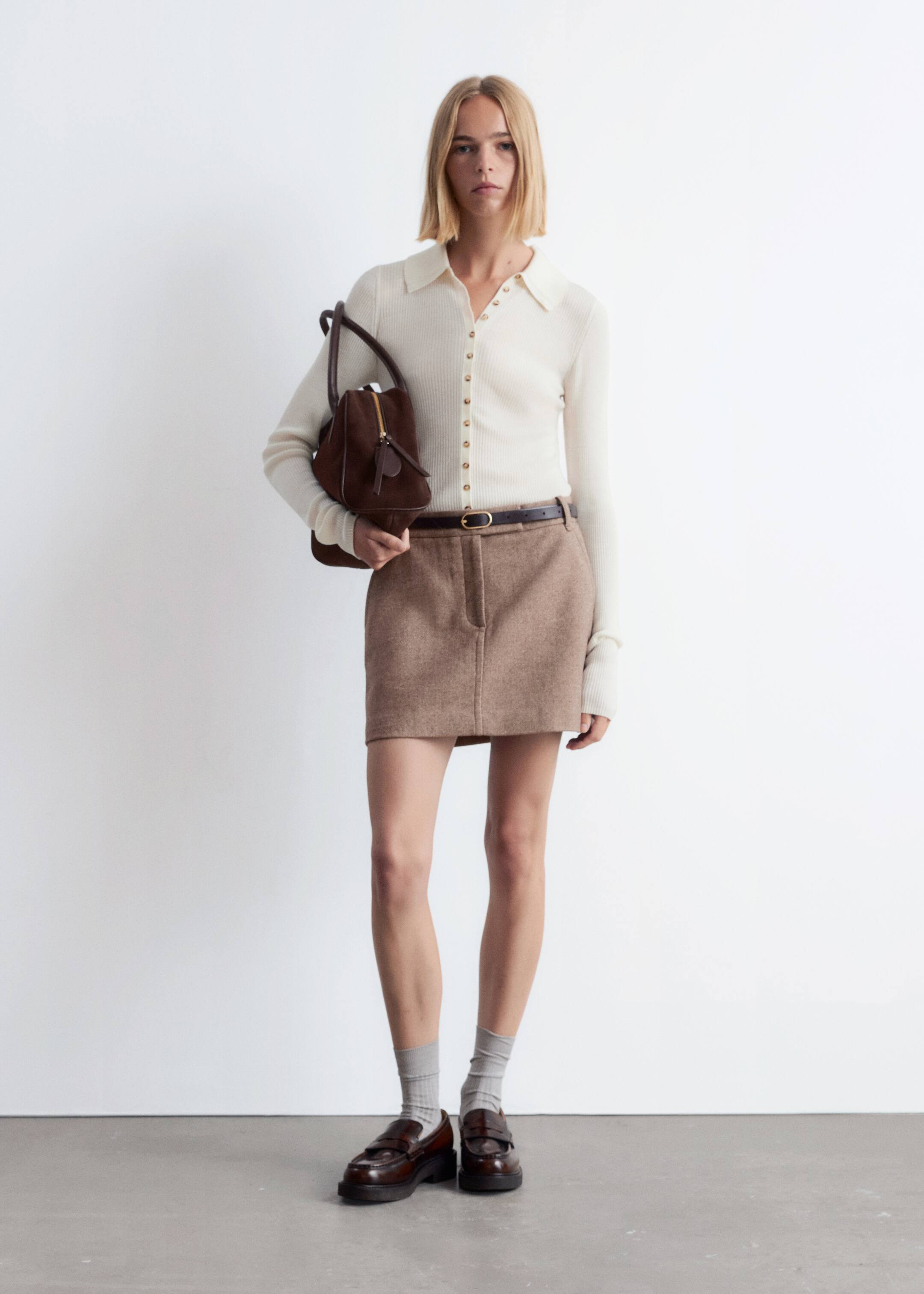 Grotere afbeelding bekijken: Een bruine, getextureerde minirok wordt van voren gepresenteerd, met een rechte silhouet en een prominente naad aan de voorkant, die boven de knie valt. Het wordt gestyled met een donkerbruine riem die door de lussen is geregen.