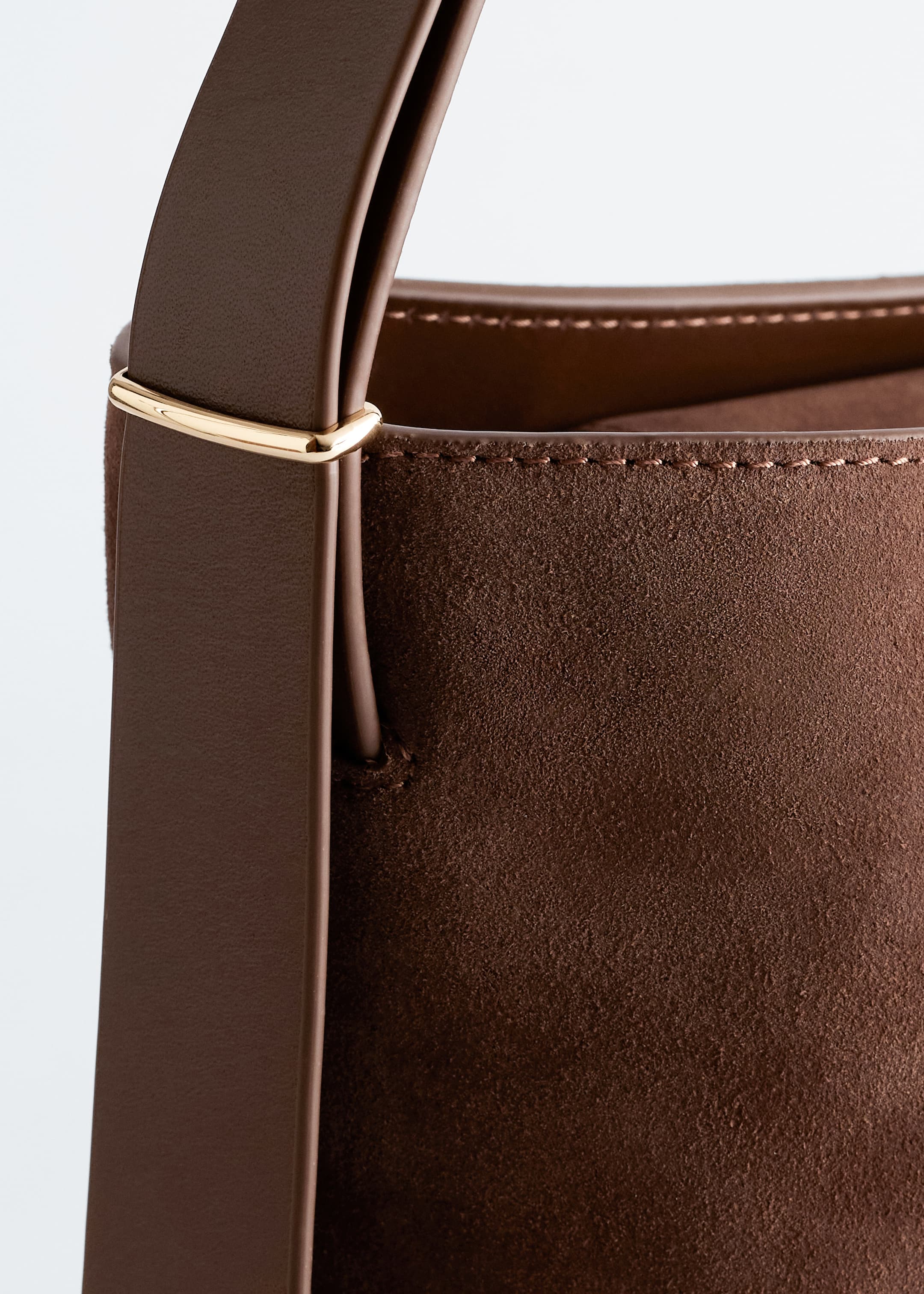 View larger image: A dark brown suede bucket bag is featuring a smooth leather shoulder strap extending upwards on the LEFT, attaching to the bag with a gold-tone metal ring, and showing visible stitching along its top edge.