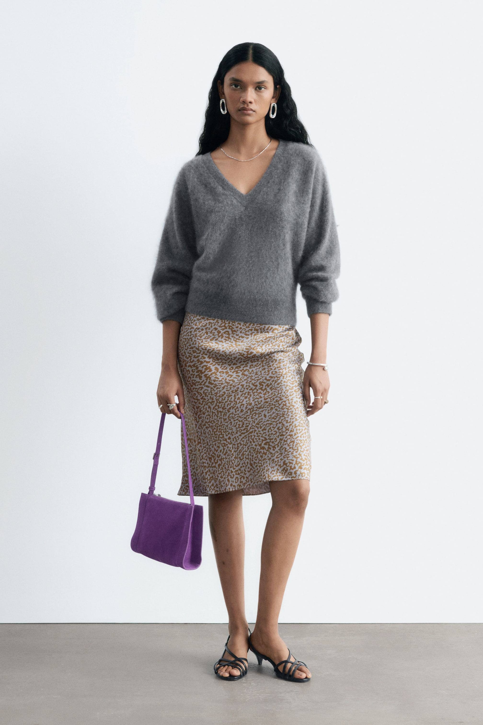 Größeres Bild anzeigen: Der Model trägt einen Satinrock mit Leopardenprint, der durch eine schmale, knielange Silhouette besticht. Der Rock ist mit einem grauen Strickpullover mit V-Ausschnitt, schwarzen Sandalen mit Riemen und einer lila Handtasche kombiniert, die in einer Hand gehalten wird.