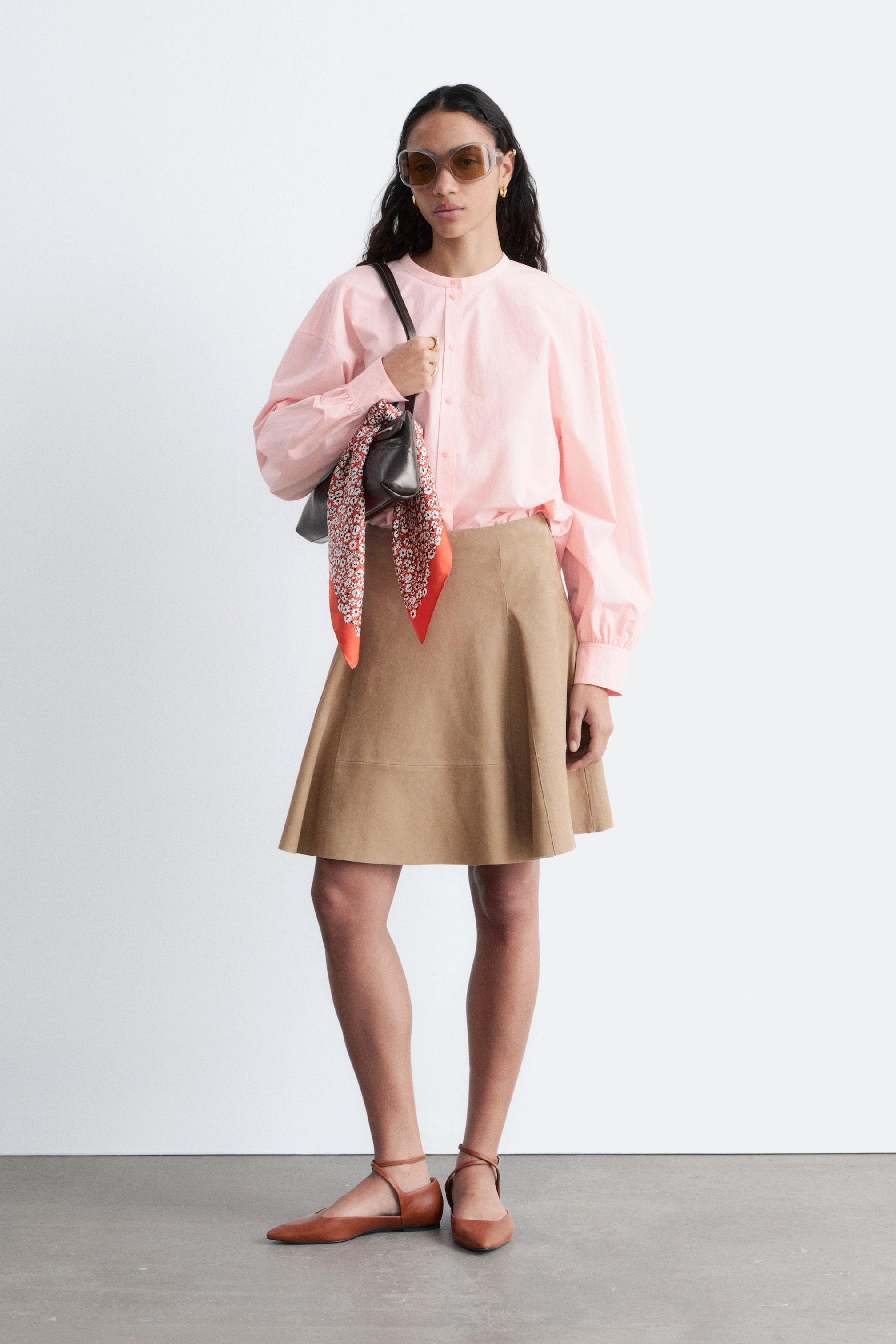 Visualizza immagine più grande: Una camicetta con collo alla coreana di colore rosa chiaro presenta maniche lunghe a sbuffo con polsini abbottonati, infilata in una gonna a trapezio color beige con cuciture a vista. Una borsa a spalla nera in stile hobo, arricchita da un piccolo foulard con motivo legato al manico, viene portata, mentre delle ballerine d'Orsay marroni a punta con cinturini alla caviglia completano il look.