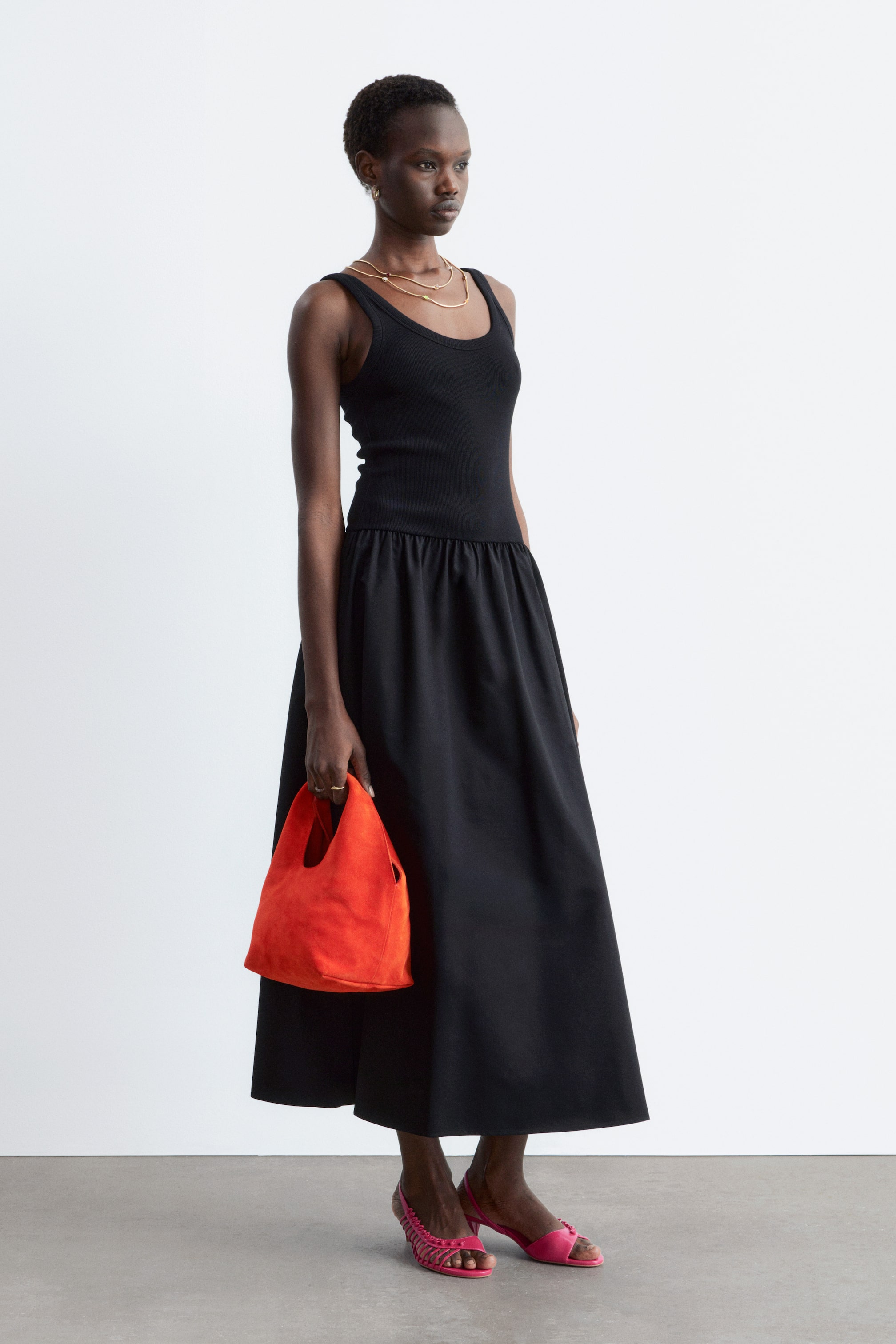 Agrandir l'image: Une robe noire présente un haut ajusté en maille côtelée avec un col rond, se prolongeant en une jupe midi ample et froncée à taille basse, et présentée sous un léger angle latéral.