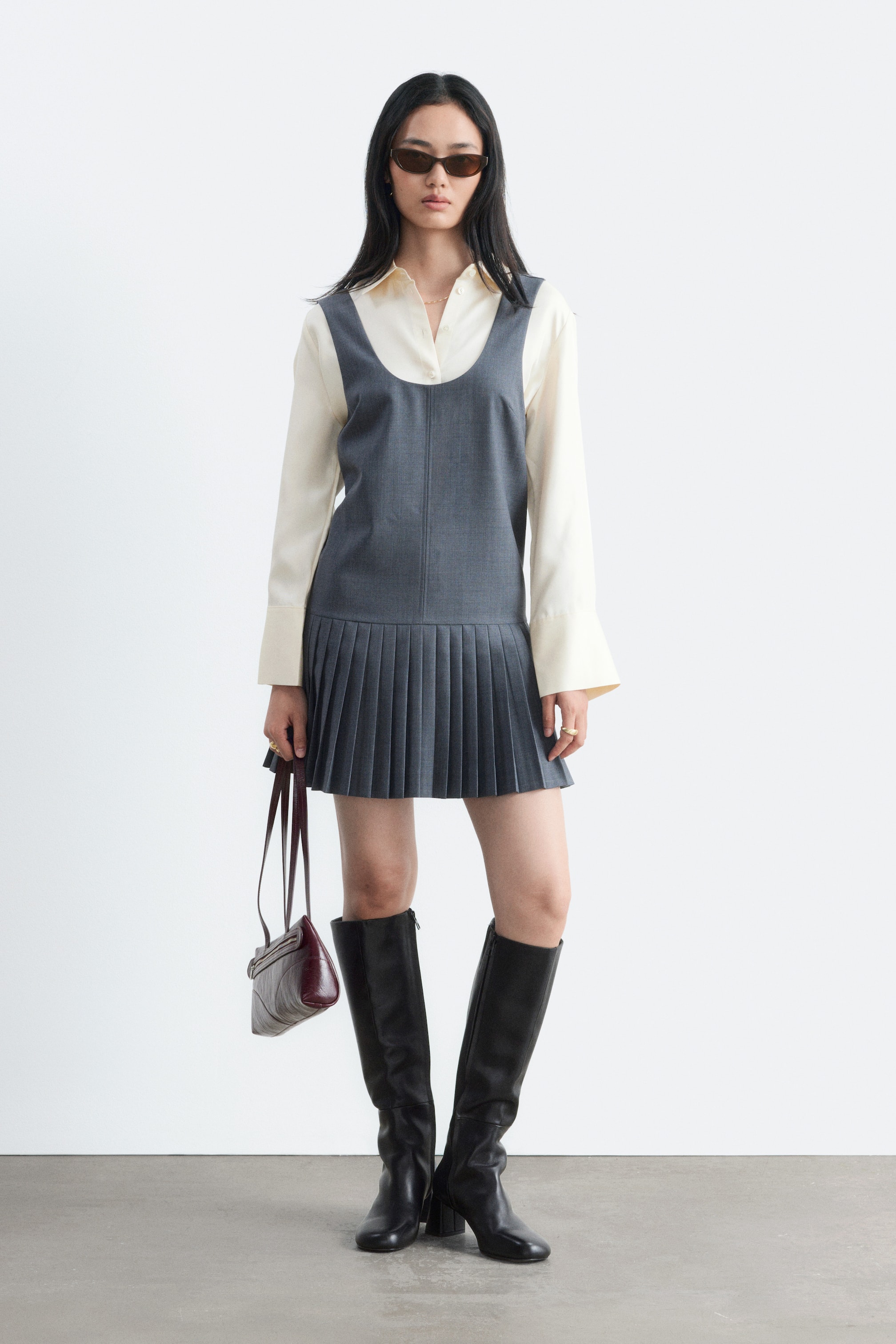 Agrandir l'image: Une robe grise courte sans manches, dotée d'un col rond et d'une jupe plissée, est portée par le mannequin. La robe est stylisée en superposition sur une chemise crème à manches longues.