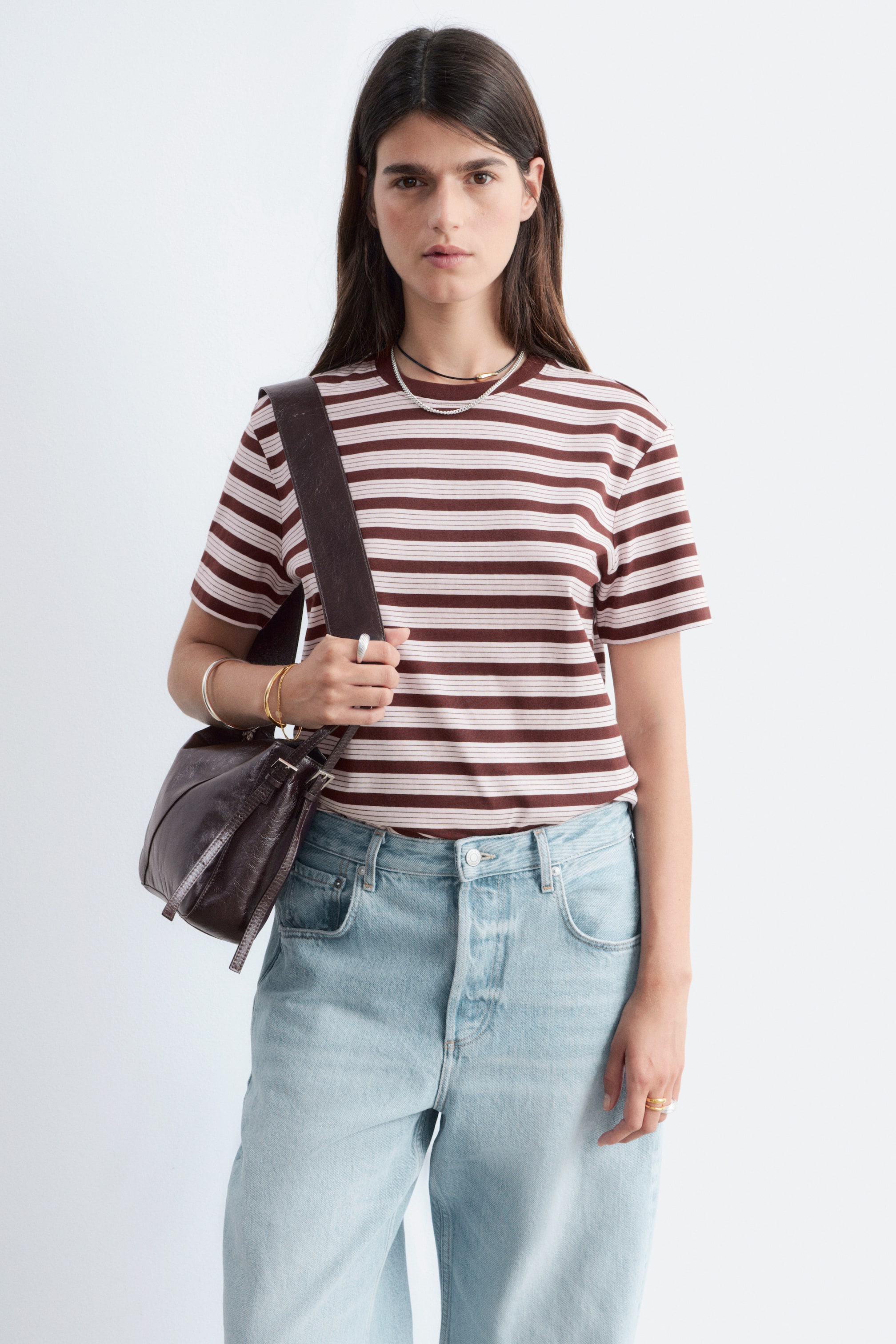 Agrandir l'image: Un t-shirt avec des rayures horizontales marron et blanc cassé, des manches courtes et un col rond est porté, vu de face. Un sac à bandoulière marron foncé traverse le t-shirt.