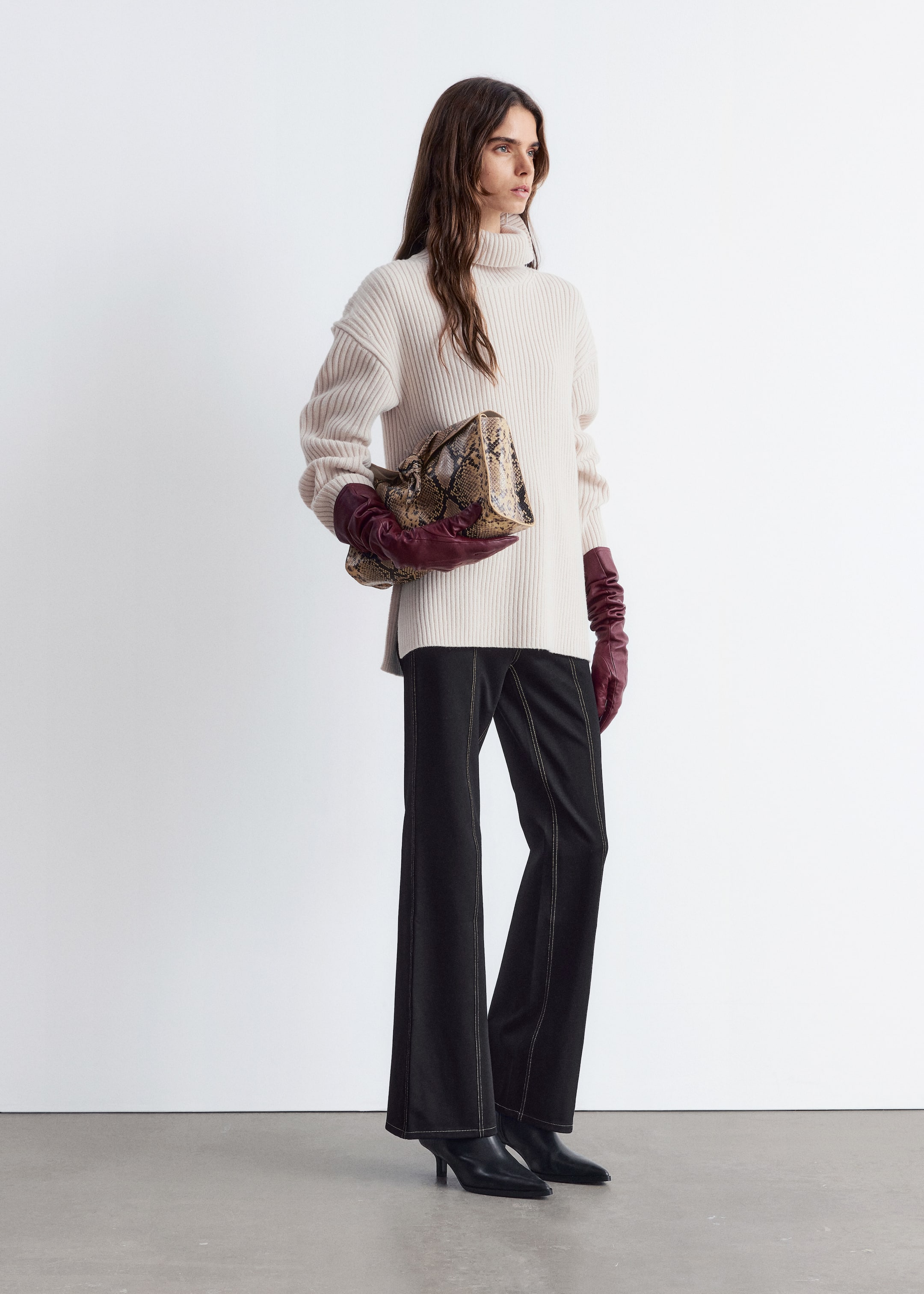 Grösseres Bild ansehen: Ein Outfit wird an einem Modell präsentiert, das leicht nach rechts geneigt ist, um Design und Passform zu zeigen. Ein cremefarbener, gerippter Rollkragenpullover mit übergroßer Passform und Seitenschlitzen wird über schwarzen Schlaghosen mit sichtbaren Kontrastnähten getragen. Der Look wird mit langen burgunderfarbenen Lederhandschuhen, einer Schlangenleder-Print-Softtasche und schwarzen spitzen Stiefeletten mit Kitten-Heels vervollständigt.