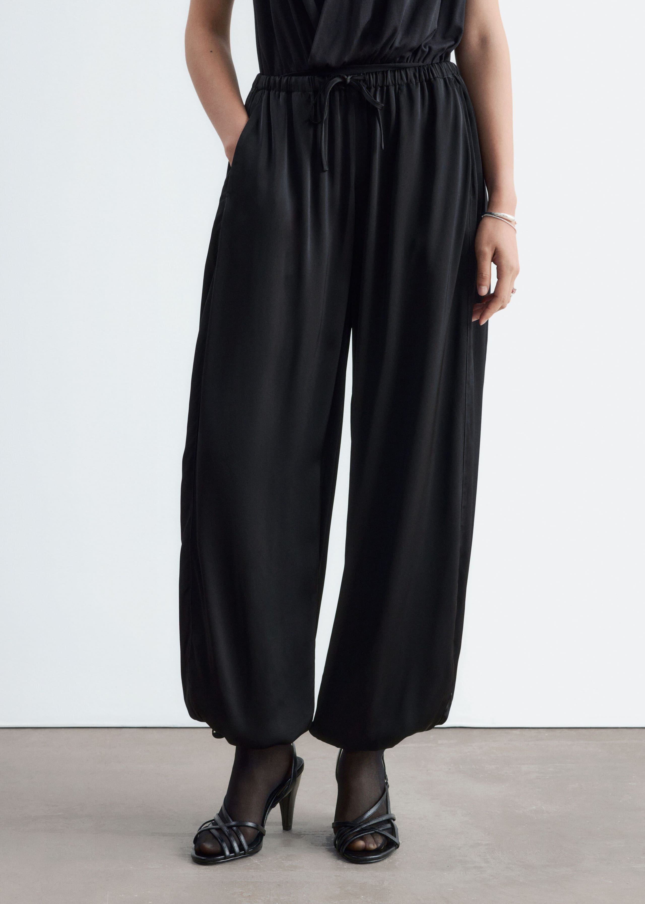 Black balloon-leg trousers made from a fluid fabric with a subtle sheen are worn by the model, who is standing with one hand in a side pocket. The trousers feature an elasticated drawstring waistband and gathered ankles, and are styled with a matching black top and strappy heeled sandals.
