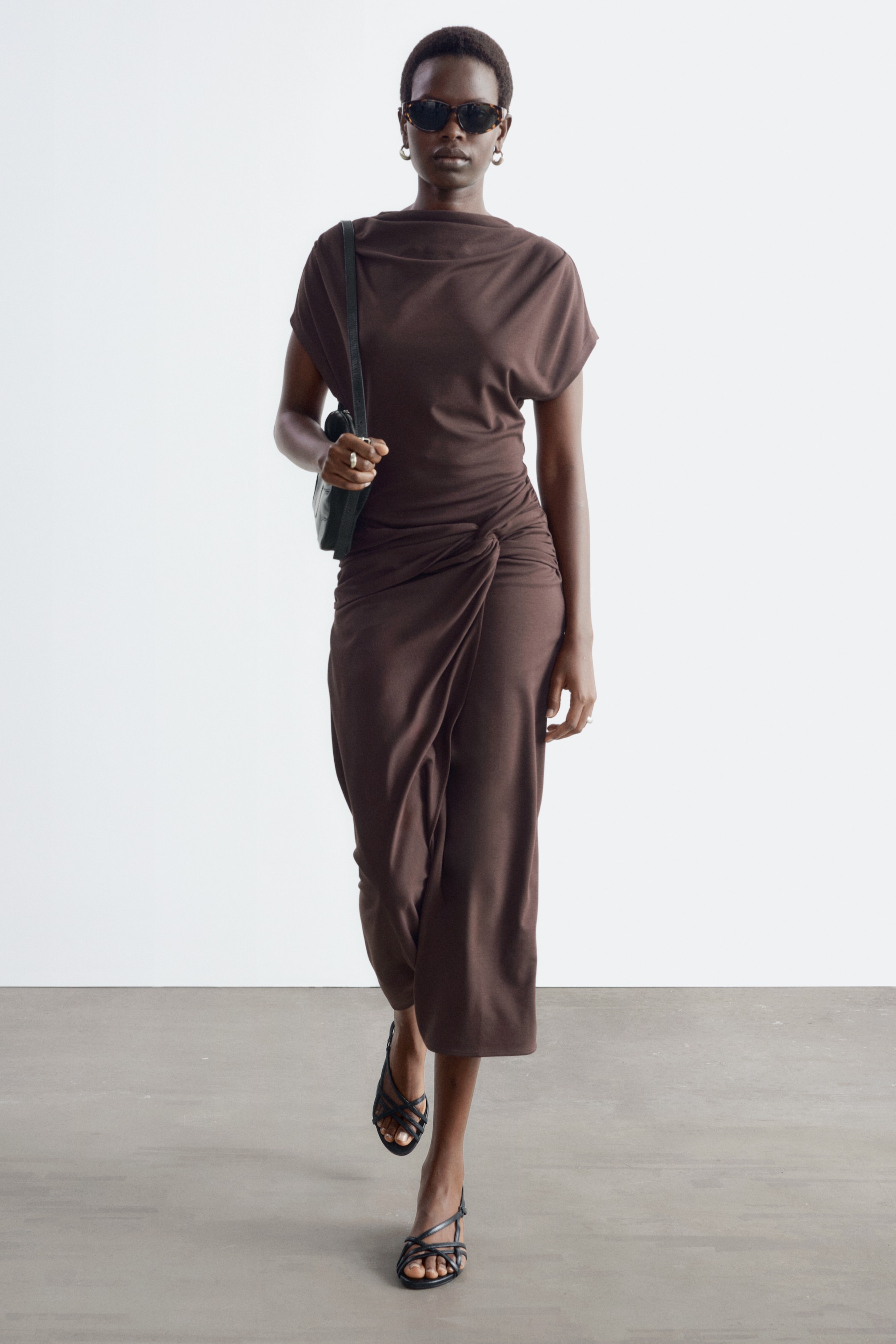 View larger image: A brown draped wrap-waist dress featuring a high draped neckline, cap sleeves, and a gathered knot detail at the waist is being worn by the model. The midi-length dress is styled with a black shoulder bag held by the strap and black strappy sandals.