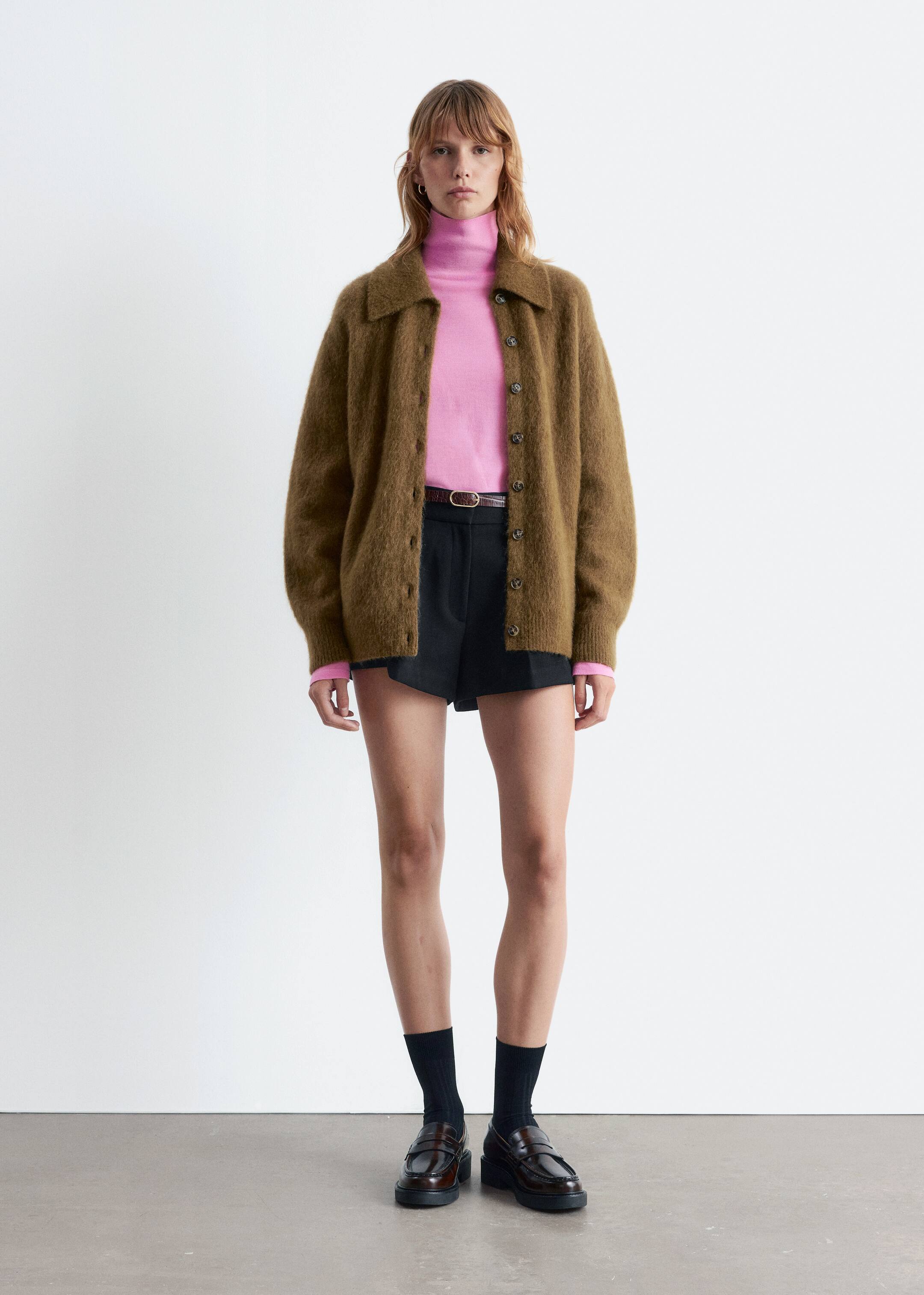 Größeres Bild ansehen: Ein ganzheitliches Outfit wird von vorne präsentiert, mit einer flauschigen moosbraunen Strickjacke mit Knopfleiste und spitzem Kragen. Ein leuchtend pinker Rollkragenpullover ist darunter geschichtet, und schwarze maßgeschneiderte Shorts werden mit einem sichtbaren braunen Gürtel gestylt. Schwarze gerippte Socken und dunkelbraune klobige Loafer vervollständigen den Look.