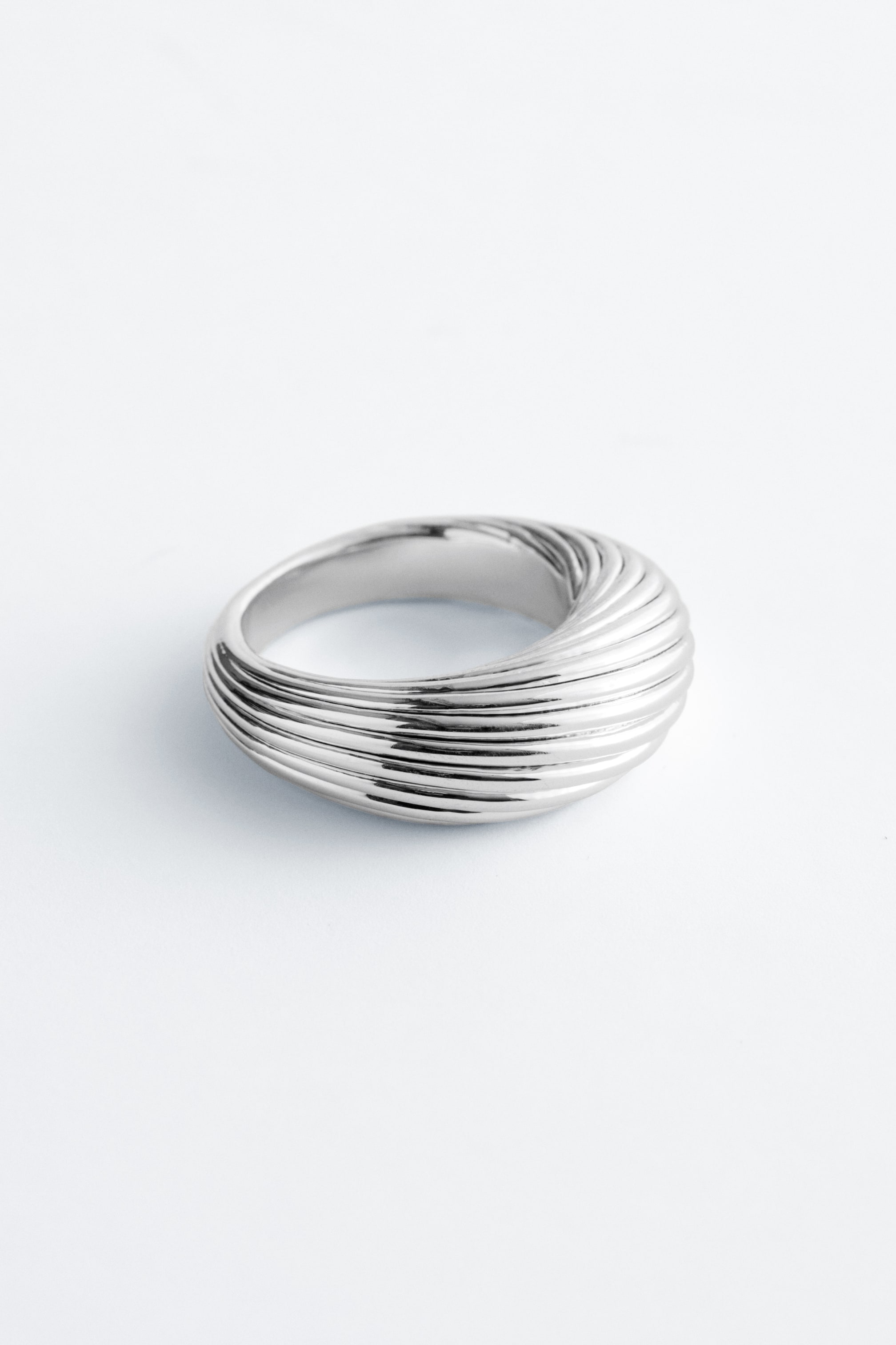 Grotere afbeelding bekijken: Een zilverkleurige ring vertoont een robuuste, sculpturale vorm. Het gepolijste oppervlak heeft een gegroefd ontwerp met parallelle, golvende ribbels die rond de voorkant wikkelen.