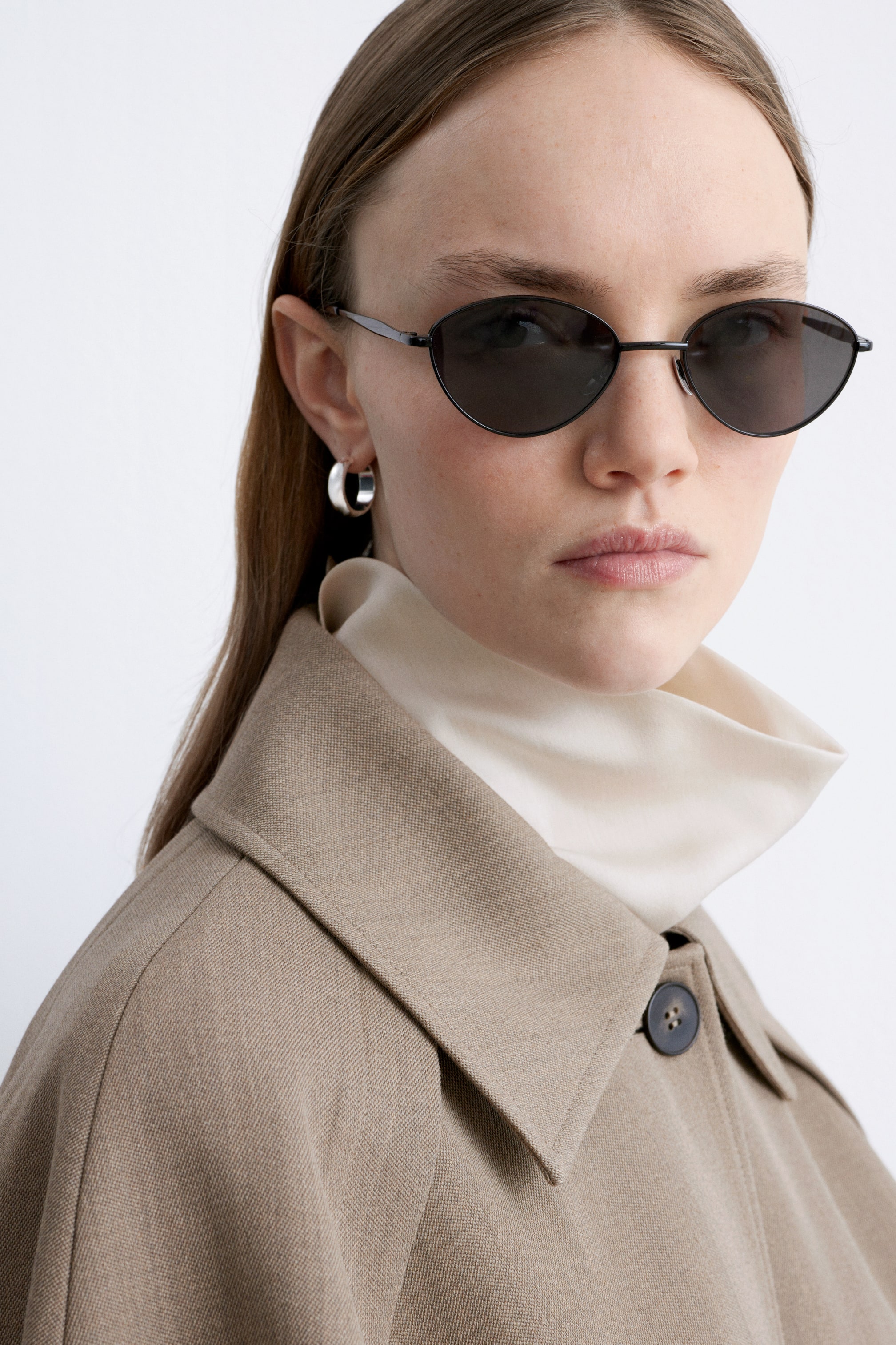 Grotere afbeelding bekijken: Ovale zonnebrillen worden gepresenteerd, met een dun zwart metalen montuur en donker getinte glazen. Ze worden gedragen op het gezicht van een persoon, gestyled met een crèmekleurige mock neck top en een beige jas met een brede puntige kraag en een donkere knoop.