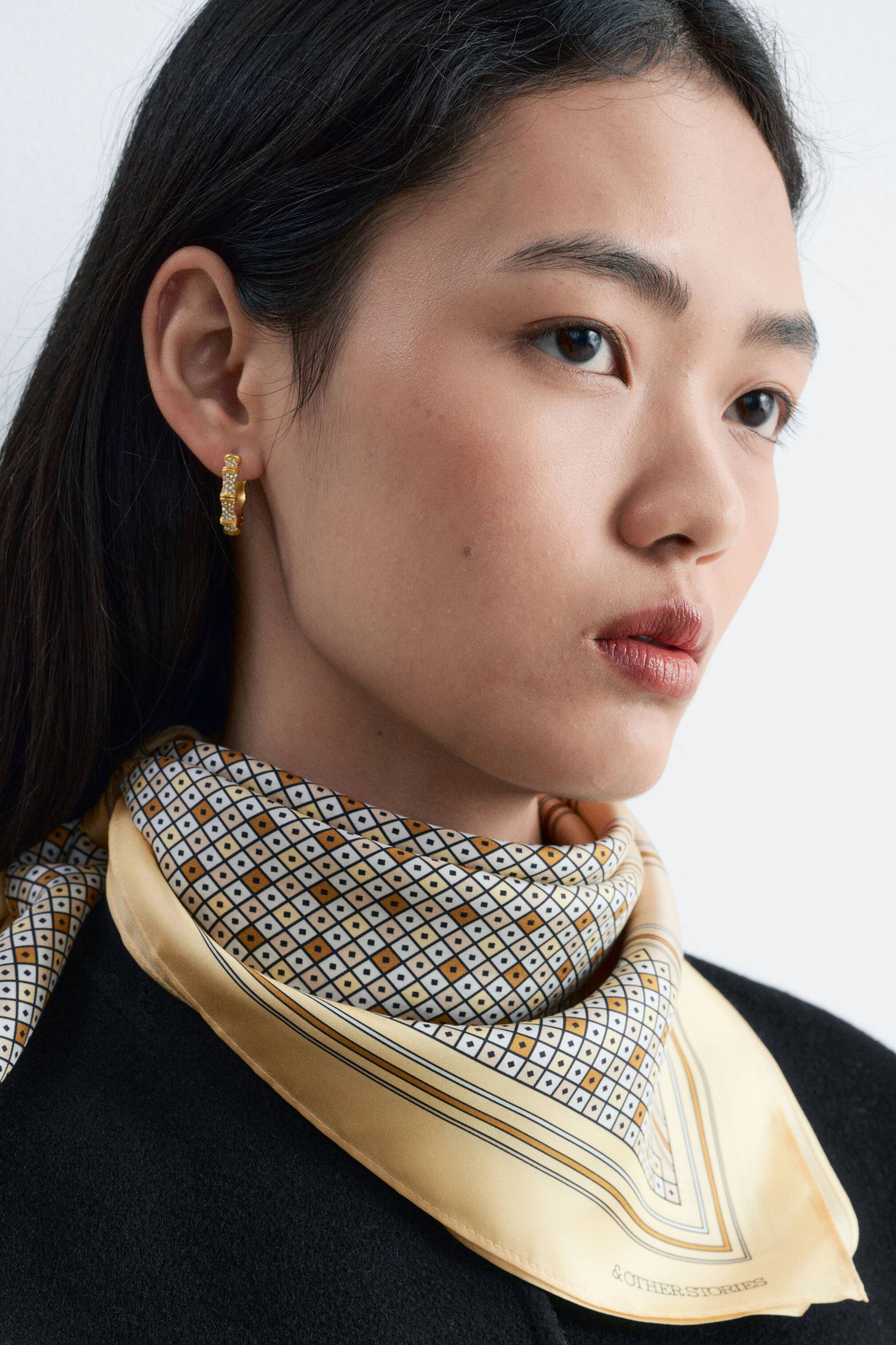 View larger image: A gold-toned hoop earring featuring a segmented, bamboo-style design with rhinestones is being worn on the model’s ear. A silk scarf with a cream and yellow geometric diamond print is also being worn tied around the model’s neck, draped over a black top.