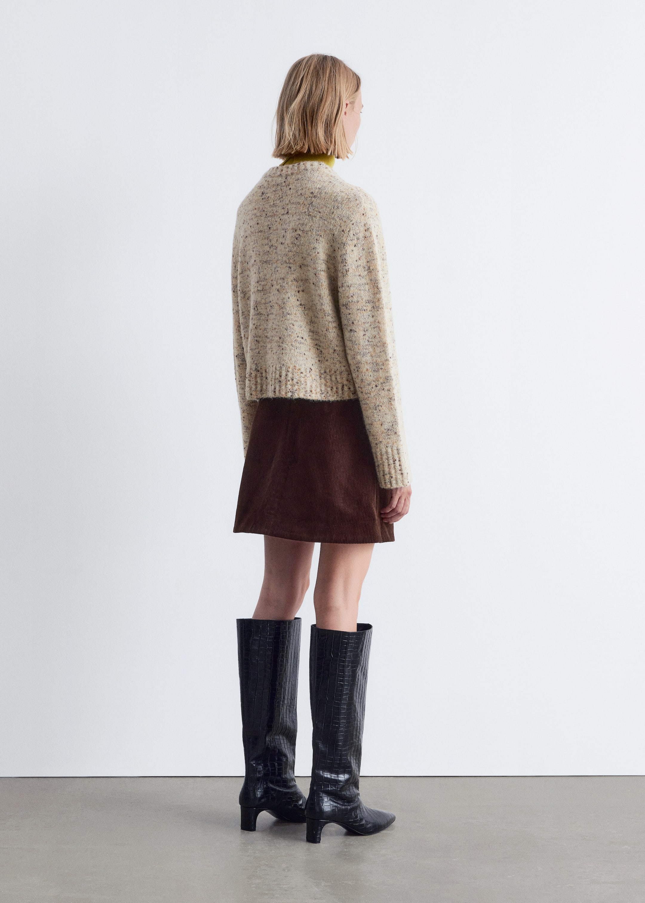 Grösseres Bild ansehen: Ein gesprenkelter cremefarbener, kurzer Pullover mit gerippten Bündchen und Saum wird über einem chartreusefarbenen Rollkragenpullover getragen, kombiniert mit einem braunen Cord-Minirock und schwarzen kniehohen Stiefeln mit Krokodilprägung und spitzen Zehen. Das komplette Ensemble wird von hinten präsentiert.