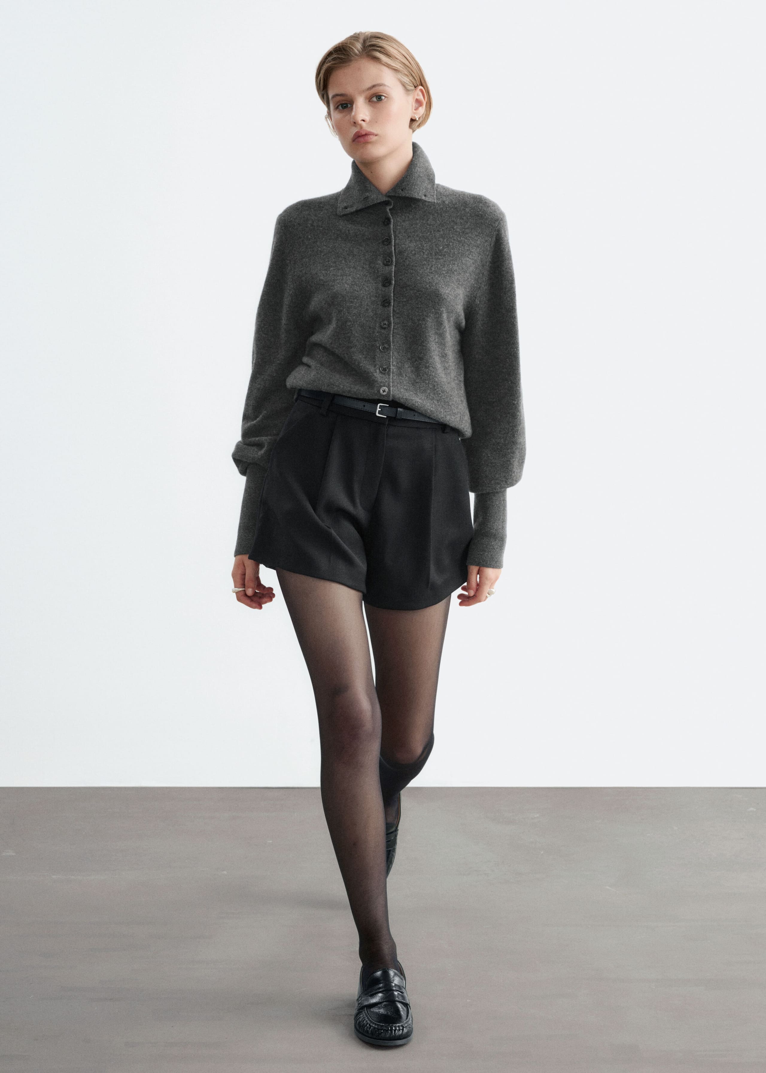 Black high-waisted shorts featuring front pleats and an A-line silhouette are worn by the model, styled with a tucked-in grey button-up cardigan and a thin black belt. The shorts are paired with sheer black tights and black loafers.