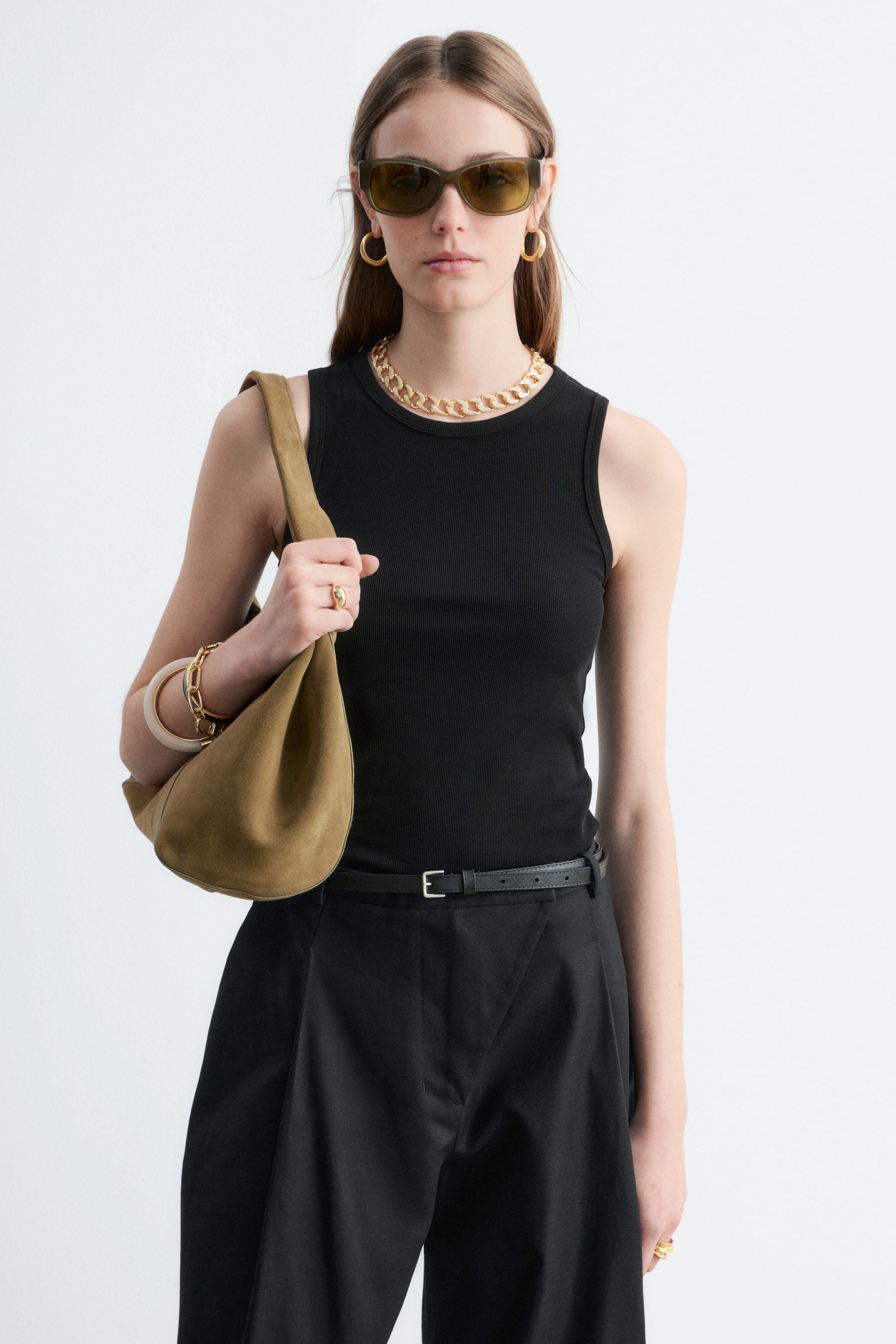 Grösseres Bild ansehen: Ein schwarzes, geripptes Jersey-Tanktop mit rundem Ausschnitt und regulärer Länge wird vom Model getragen. Das eng anliegende Oberteil ist in schwarze Hosen gesteckt, mit einer goldenen Kette, die auf dem Ausschnitt ruht.