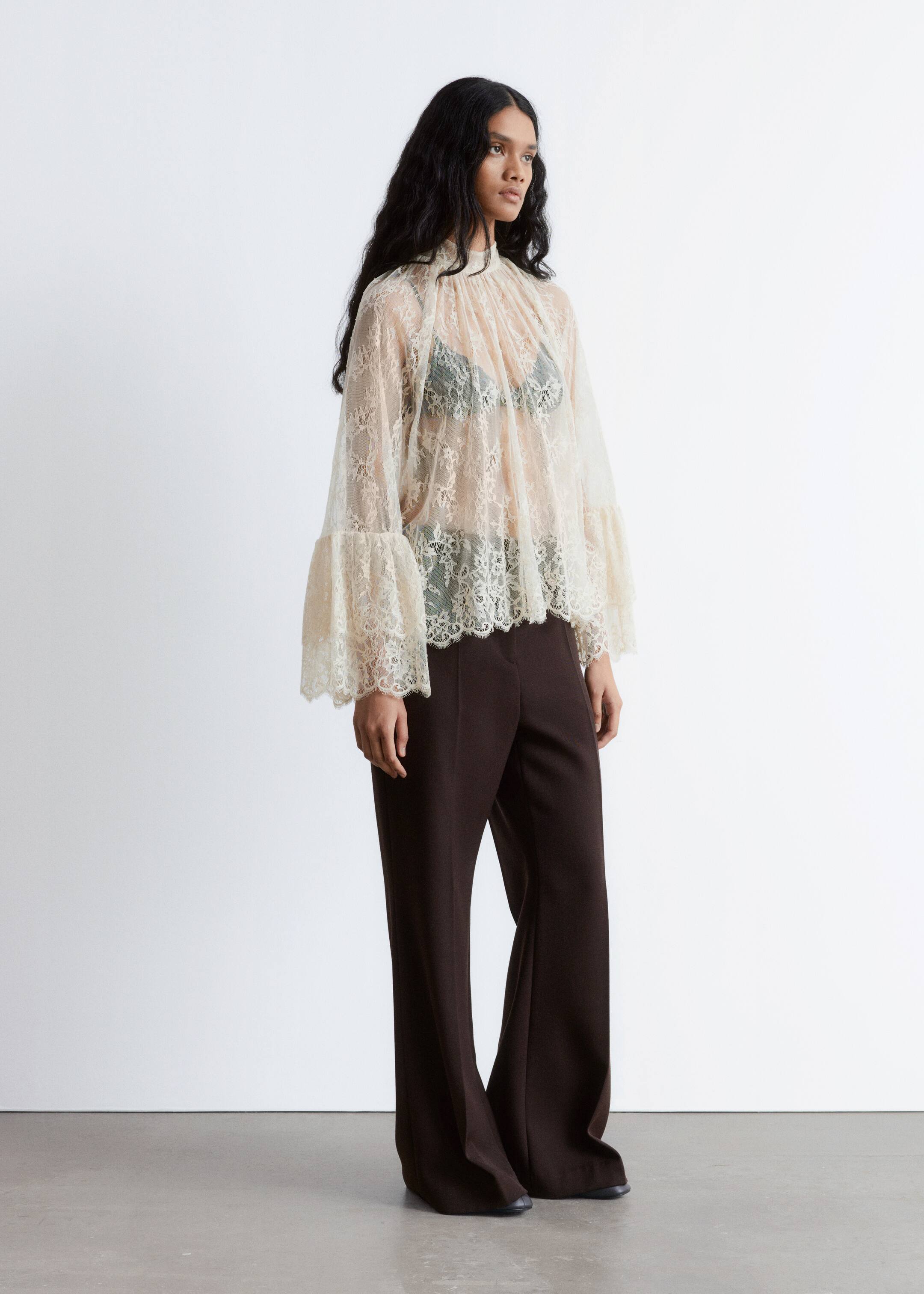 Agrandir l'image: Une blouse en dentelle légère et de couleur claire, avec un col montant à volants, des manches évasées à étages avec des poignets festonnés, et un ourlet festonné, est présentée sous un angle de 3/4, révélant un soutien-gorge foncé en dessous. Elle est assortie à un pantalon large taille haute de couleur brun foncé qui tombe jusqu'au sol.