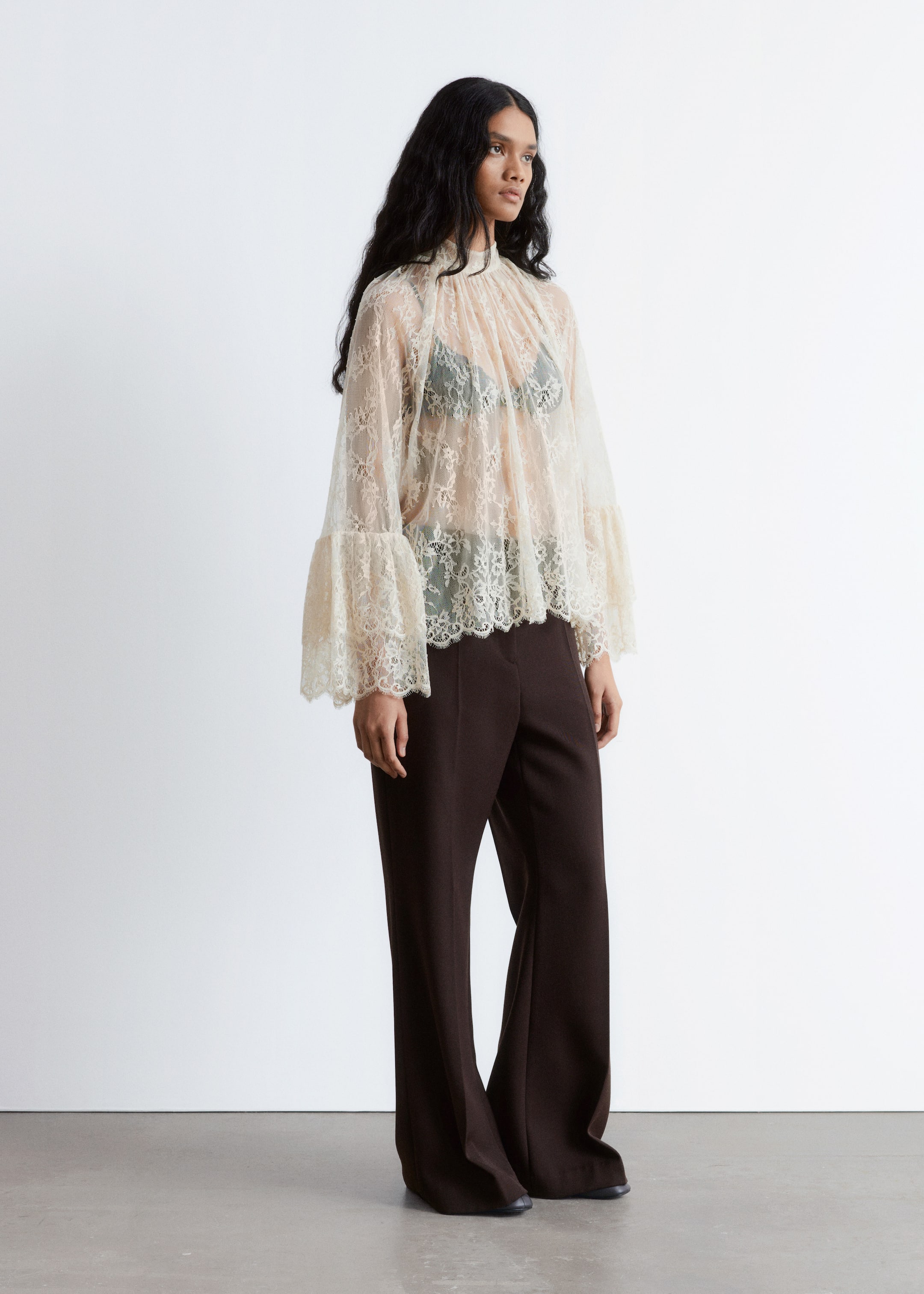 Visualizza immagine più grande: Una blusa di pizzo trasparente di colore chiaro, caratterizzata da un collo alto arricciato, maniche a campana a strati con polsini smerlati e un orlo smerlato, viene presentata da un'angolazione di 3/4, rivelando un reggiseno scuro sotto. Viene abbinata a pantaloni a vita alta, a gamba larga, di colore marrone scuro che cadono fino al pavimento.