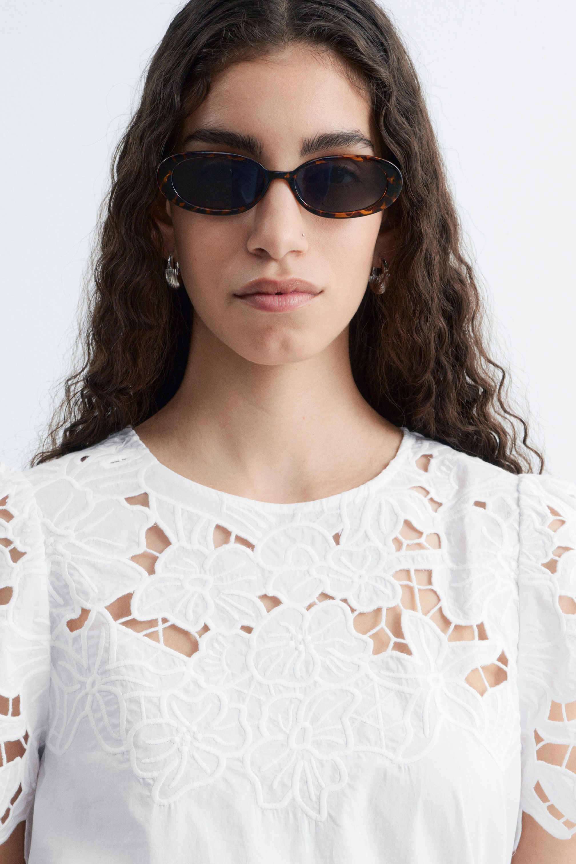 Ver imagen más grande: Una blusa de algodón blanco y crujiente está siendo llevada por la modelo. Presenta un escote redondo, mangas cortas abullonadas y un intrincado bordado floral blanco con detalles calados en el pecho y los hombros.