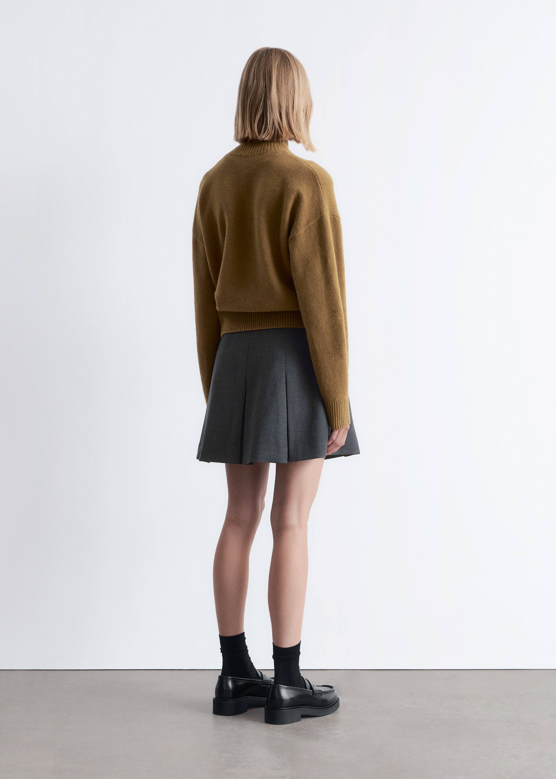 Agrandir l'image: Un pull marron moutarde, ample, est présenté de dos, avec un col montant et des poignets et un ourlet côtelés. Une mini-jupe plissée gris foncé est visible sous le pull, stylée avec des mocassins noirs et des chaussettes noires.