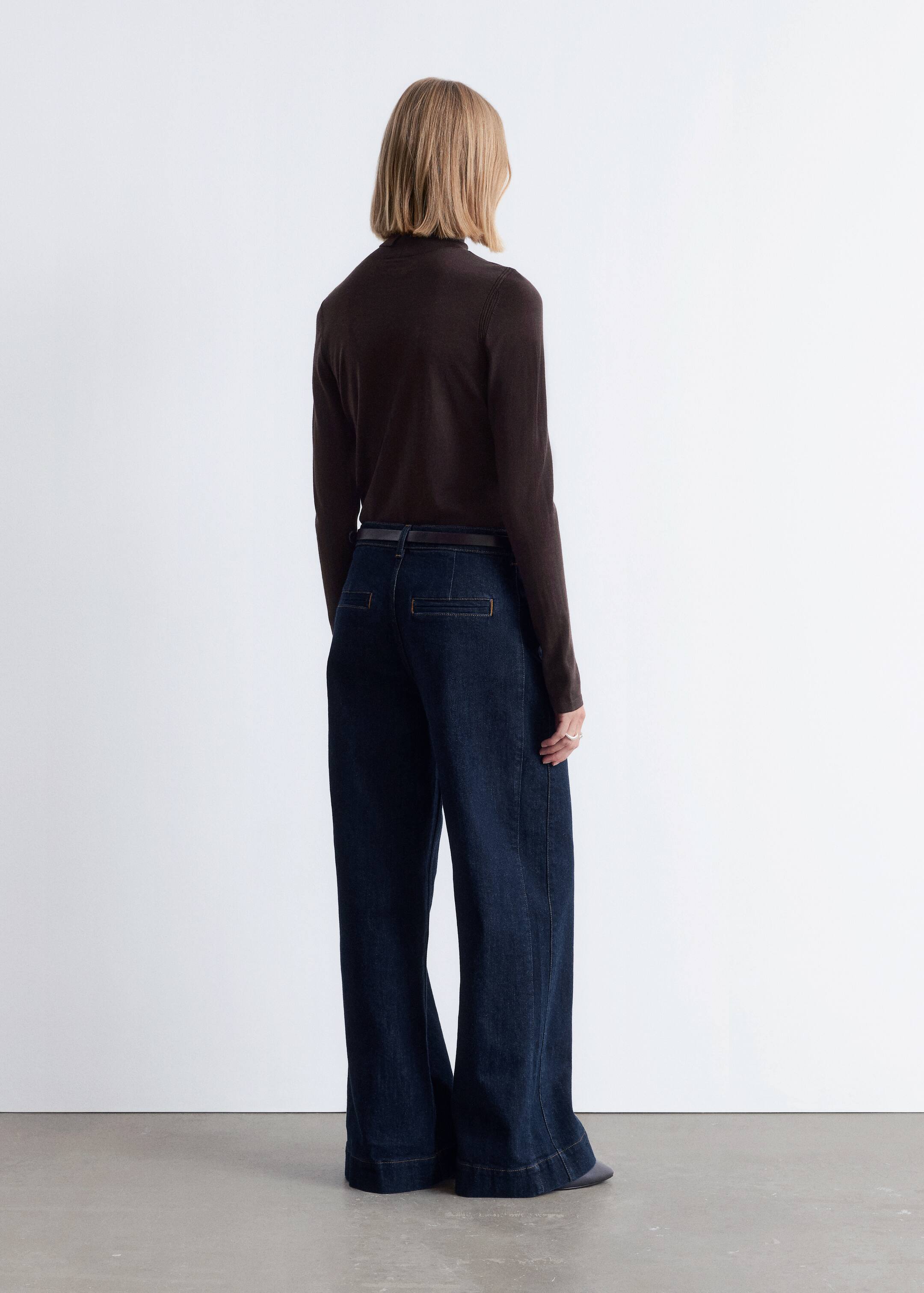 Visa större bild: Mörka tvättade jeans med vida ben visas från en bakre, något vinklad perspektiv, med synliga ljusa kontrastsömmar, platta rektangulära bakfickor och en bred fåll. Ett mörkt bälte bärs genom bältesöglorna.