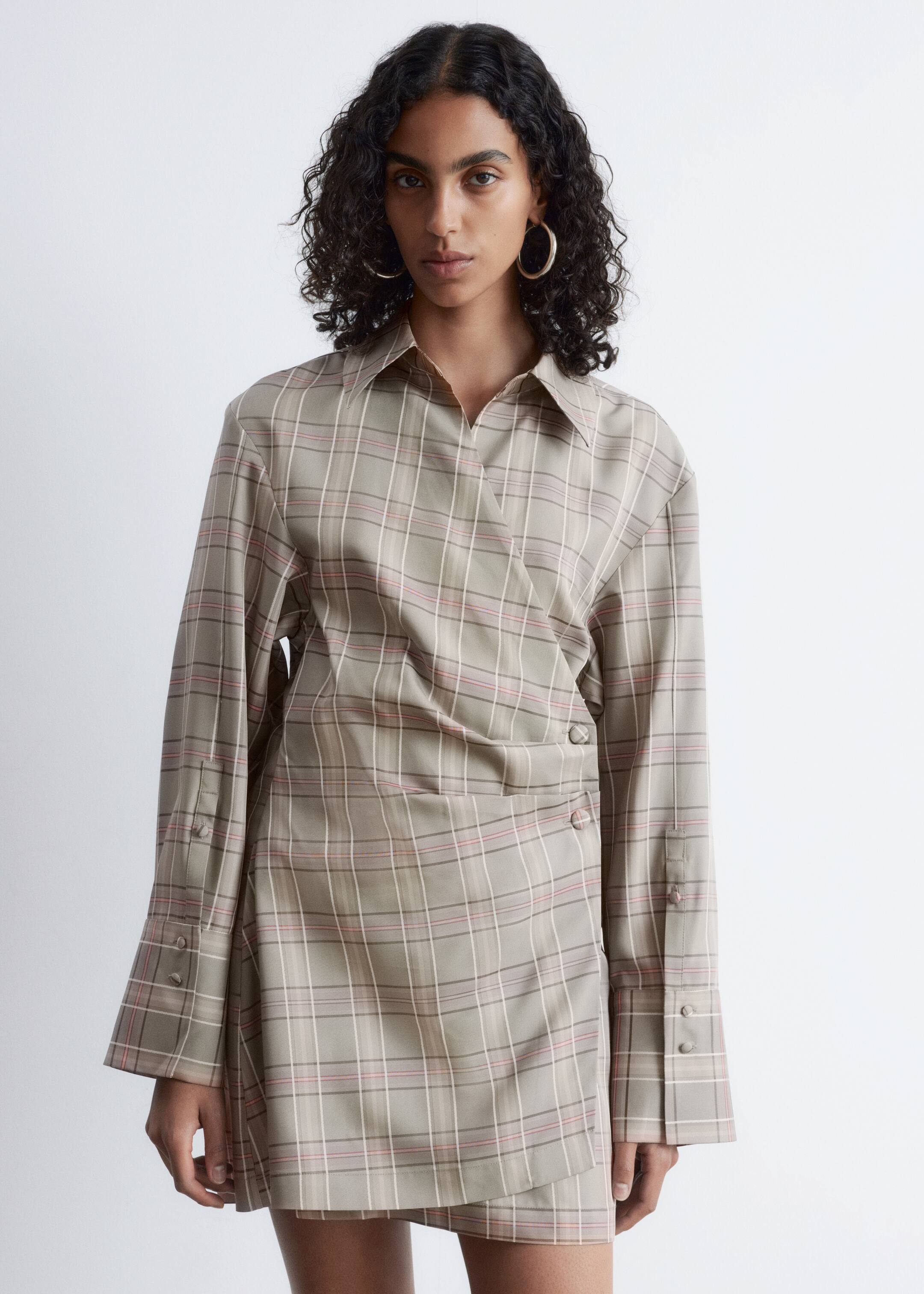 Agrandir l'image: Une robe portefeuille à carreaux portée face avant présente une base beige avec un motif à carreaux constitué de fines lignes blanches et rouges, un col V, des manches longues avec poignets à deux boutons, et une fermeture portefeuille à l'avant sécurisée par deux boutons visibles sur le côté.