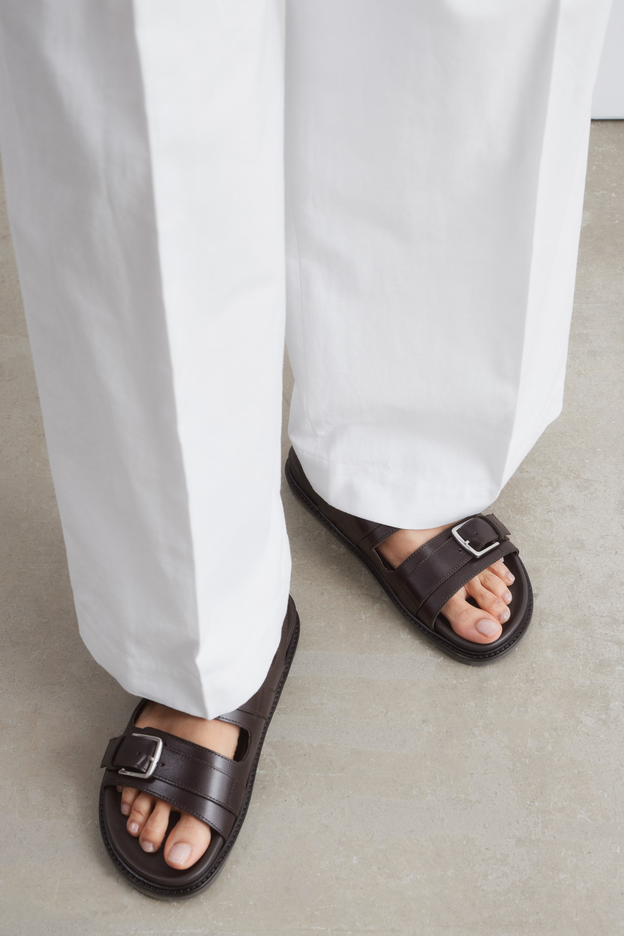 Visa större bild: Mörkbruna lädersandaler med öppen tå och två spännen över ovandelen bärs av modellen. En platt yttersula är synlig, och vita byxor täcker delvis sandalerna.