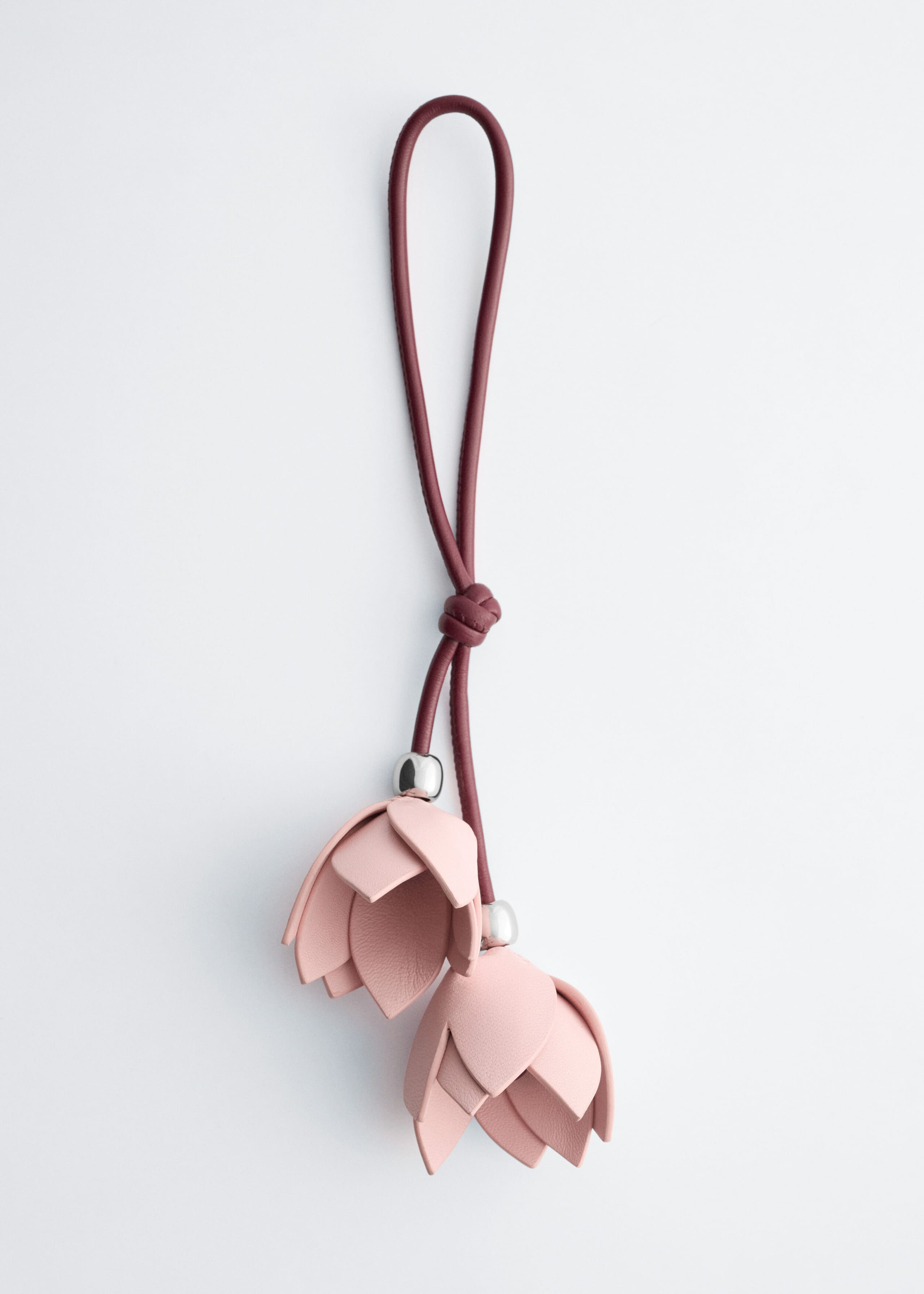 A floral bag charm hangs vertically, featuring a looped burgundy leather strap secured with a central knot. Two light pink, tulip-shaped leather flowers dangle from the ends of the strap at slightly different lengths, each accented with a polished silver-tone metal bead at its base.