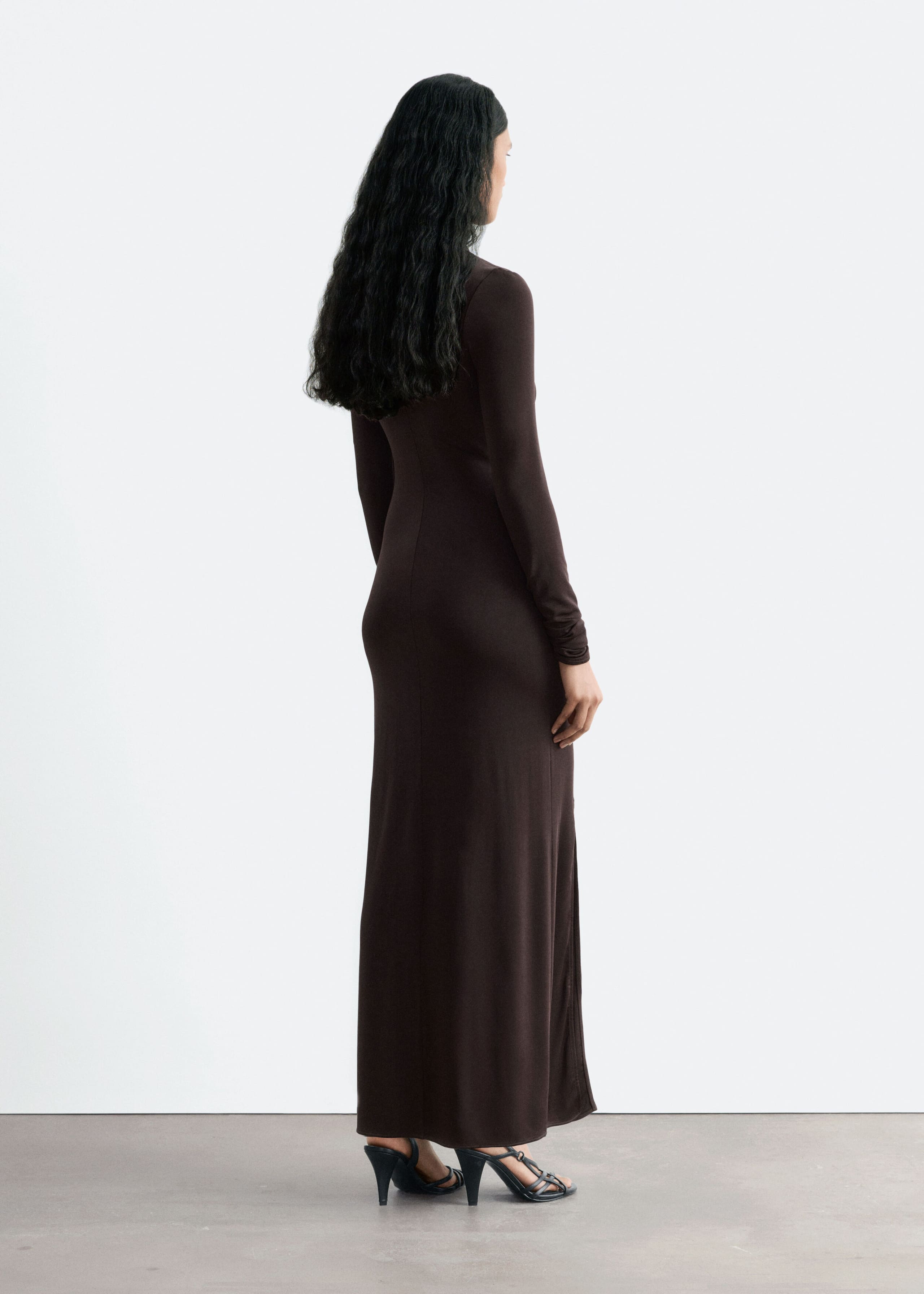 A long-sleeved, dark brown jersey maxi dress with a slim fit is being worn by the model. The model is standing turned away to show the back profile of the dress, which is styled with black strappy heeled sandals.