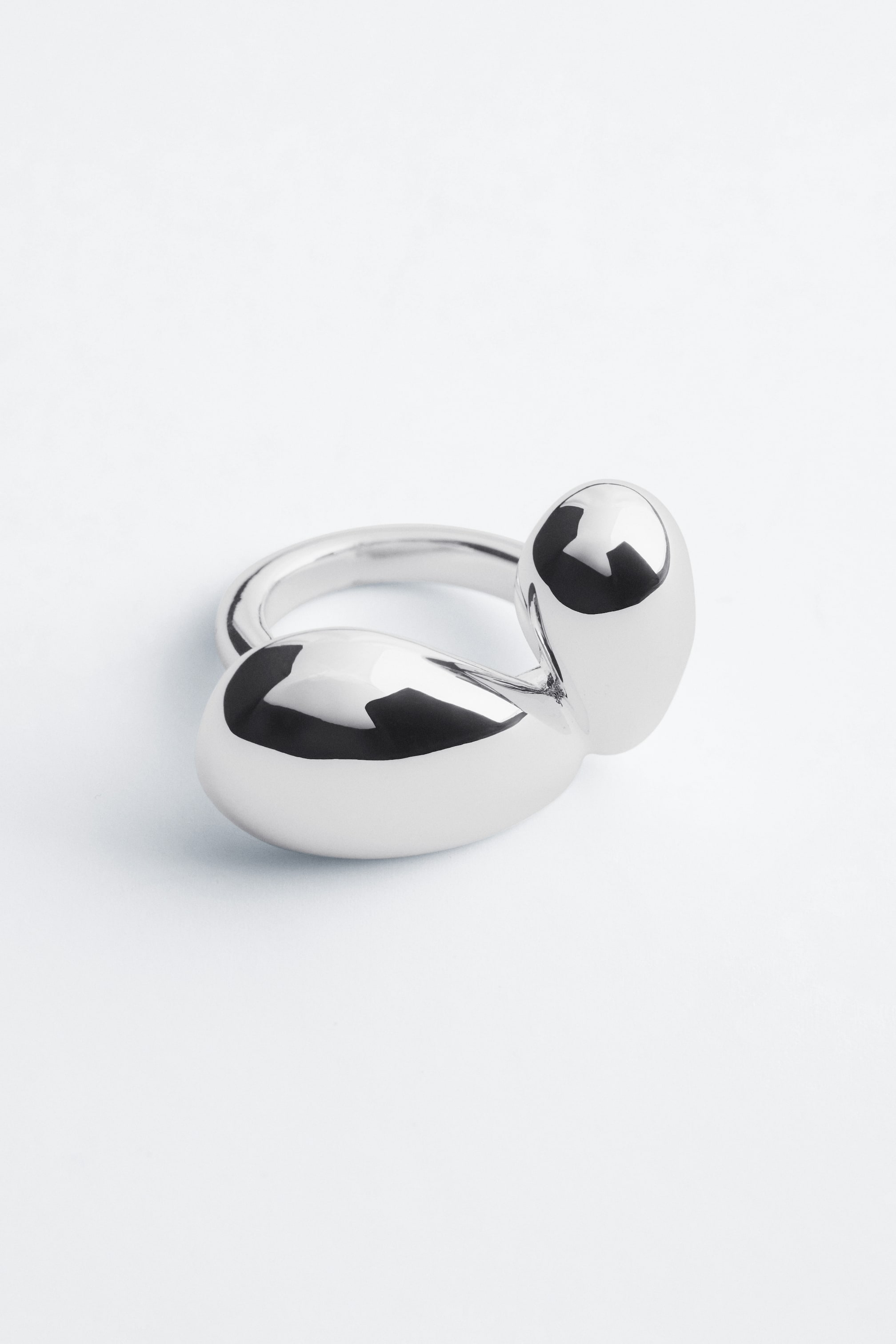 Größeres Bild ansehen: Ein polierter, silberfarbener Ring zeigt ein skulpturales Design, das von zwei fließenden, voluminösen Formen geprägt ist, die sich aus einem glatten Ringband nach oben wölben. Der Ring liegt leicht schräg, um seine reflektierende, organische Form hervorzuheben.
