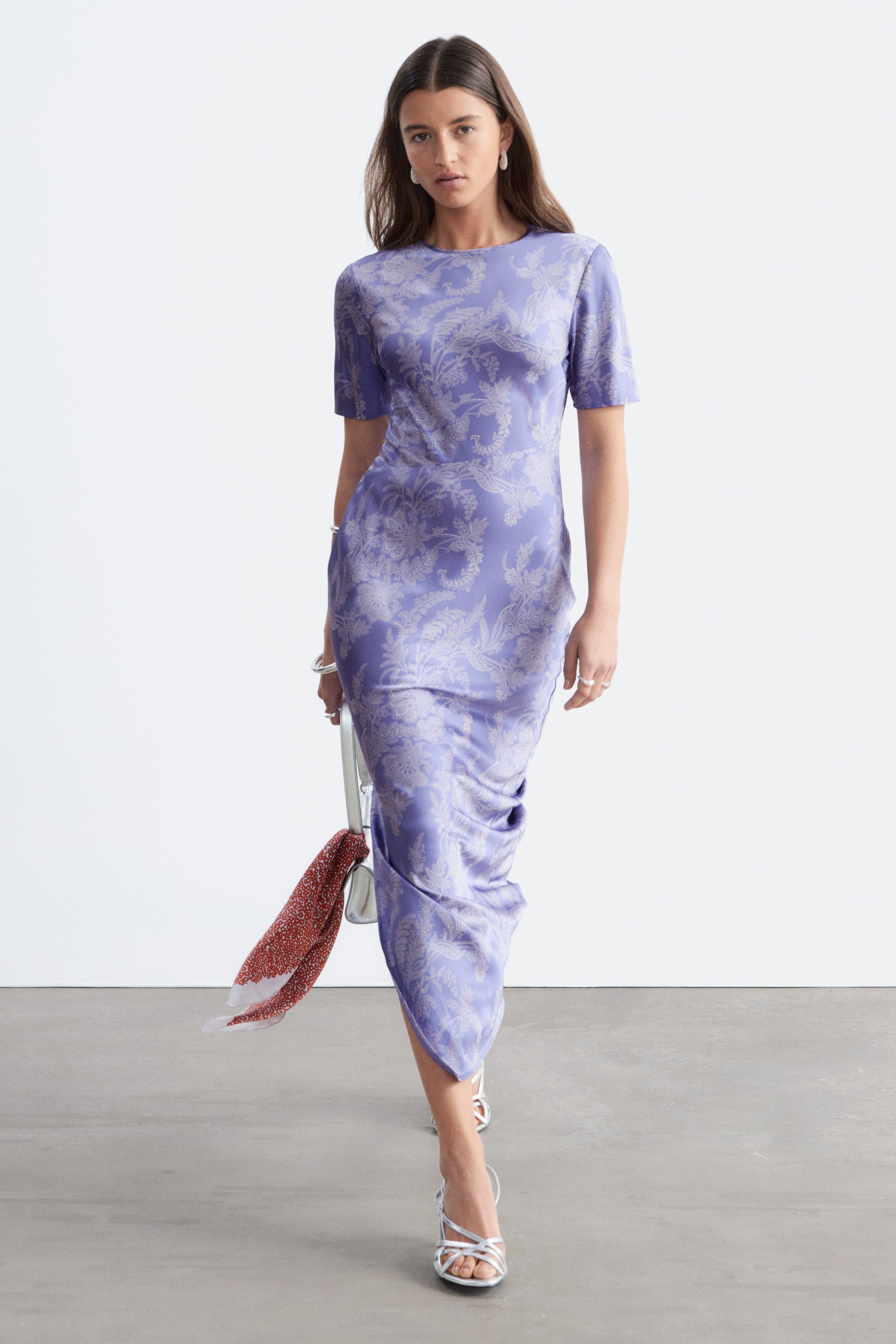 Se større billede: En lavendelfarvet midikjole med et lysere paisley-inspireret blomsterprint over det hele præsenteres fra en frontvinkel. Den har en rund halsudskæring og korte ærmer, hvor det kropsnære stof blødt draperer ned ad benene. Kjolen styles med en hvid håndtaske, der har et rødt mønstret tørklæde bundet til stroppen, samt sølvfarvede sandaler med remme.