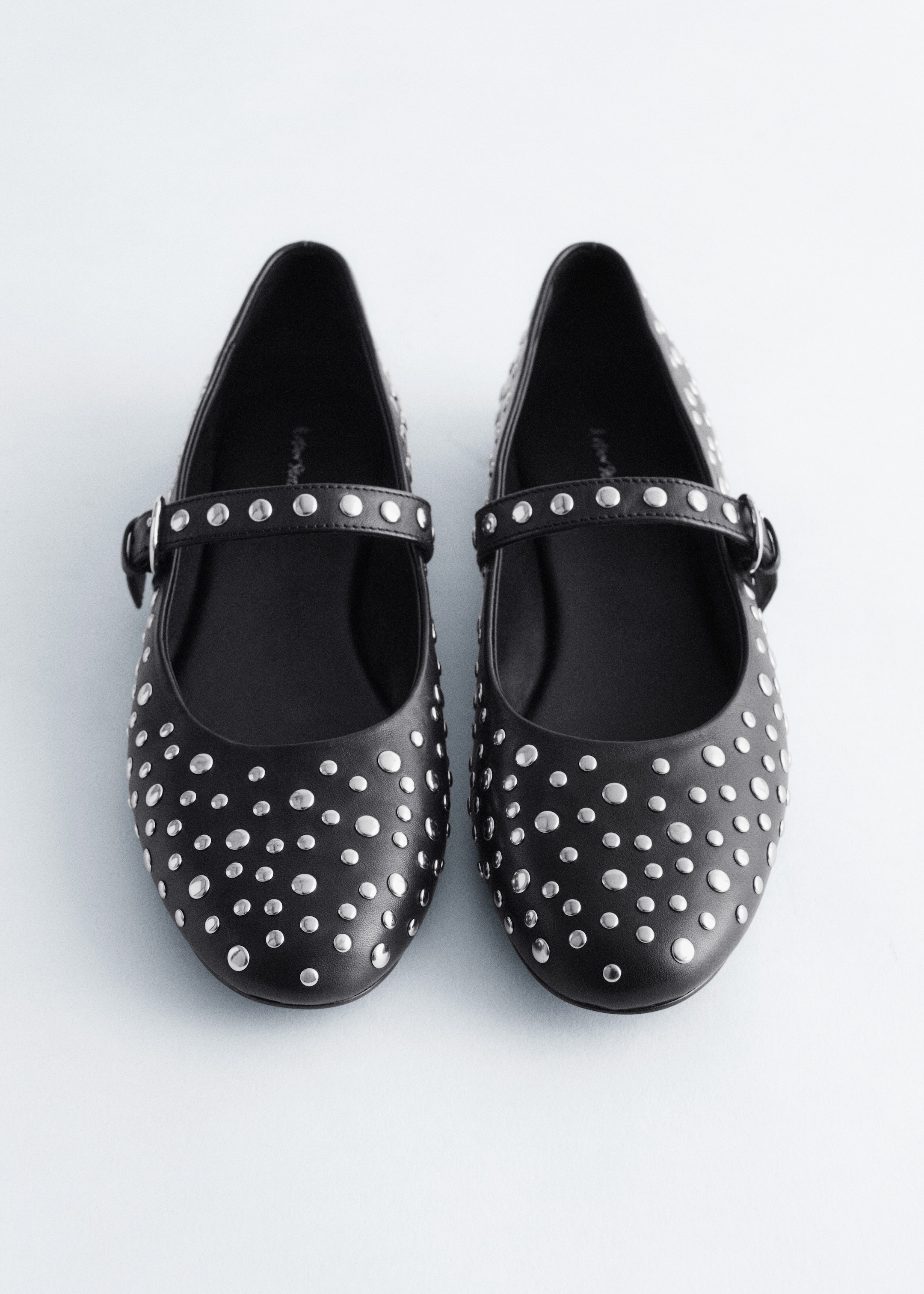 View larger image: A pair of black leather Mary Jane ballet flats is being presented from the front. Each flat is featuring allover silver-toned metal studs and a strap across the vamp that is secured by a tonal black buckle.