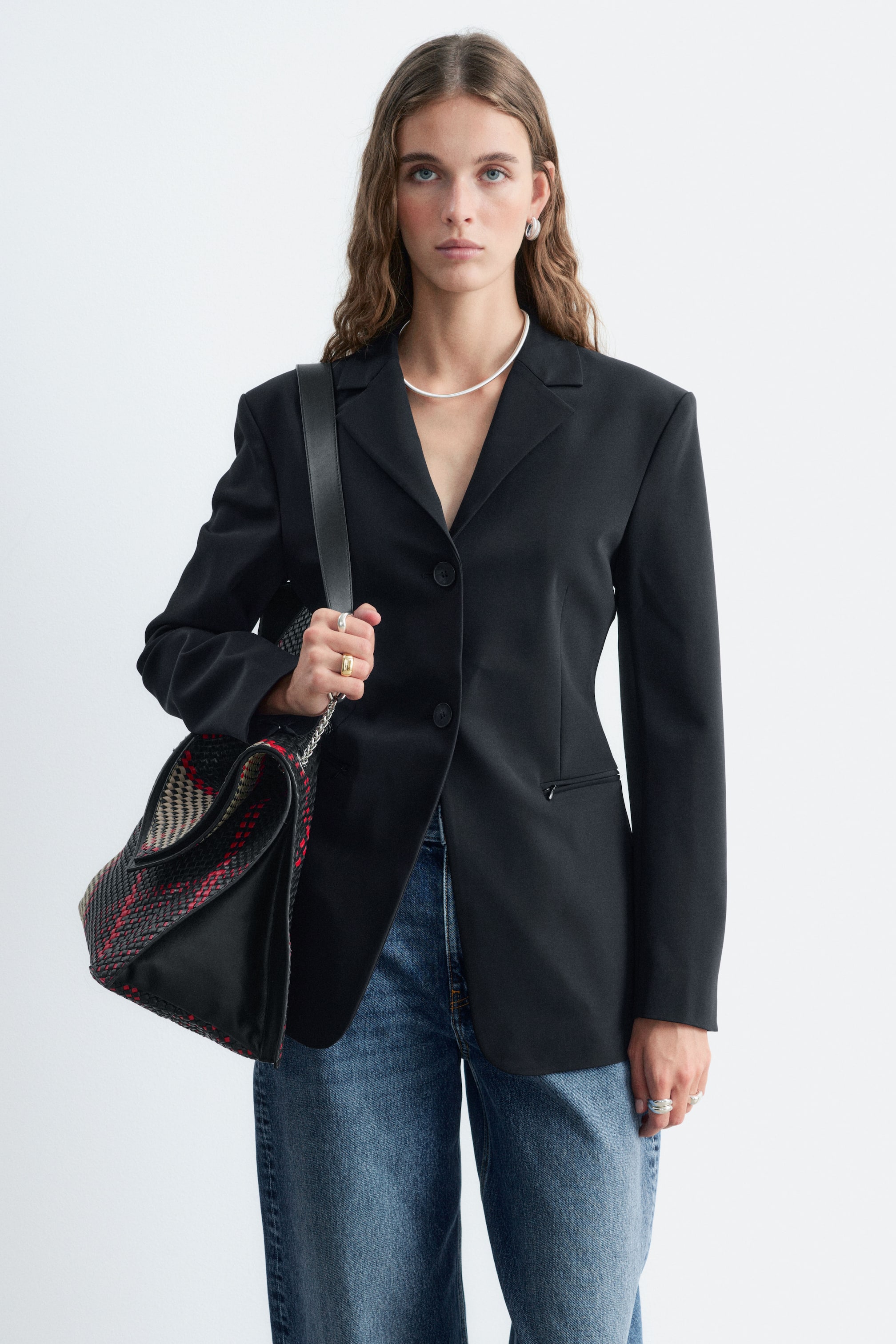 View larger image: A black tailored blazer featuring a fitted waist, notch lapels, and a two-button fastening is being worn by the model. The blazer is styled with blue jeans, with the model holding the strap of a patterned shoulder bag over one shoulder.