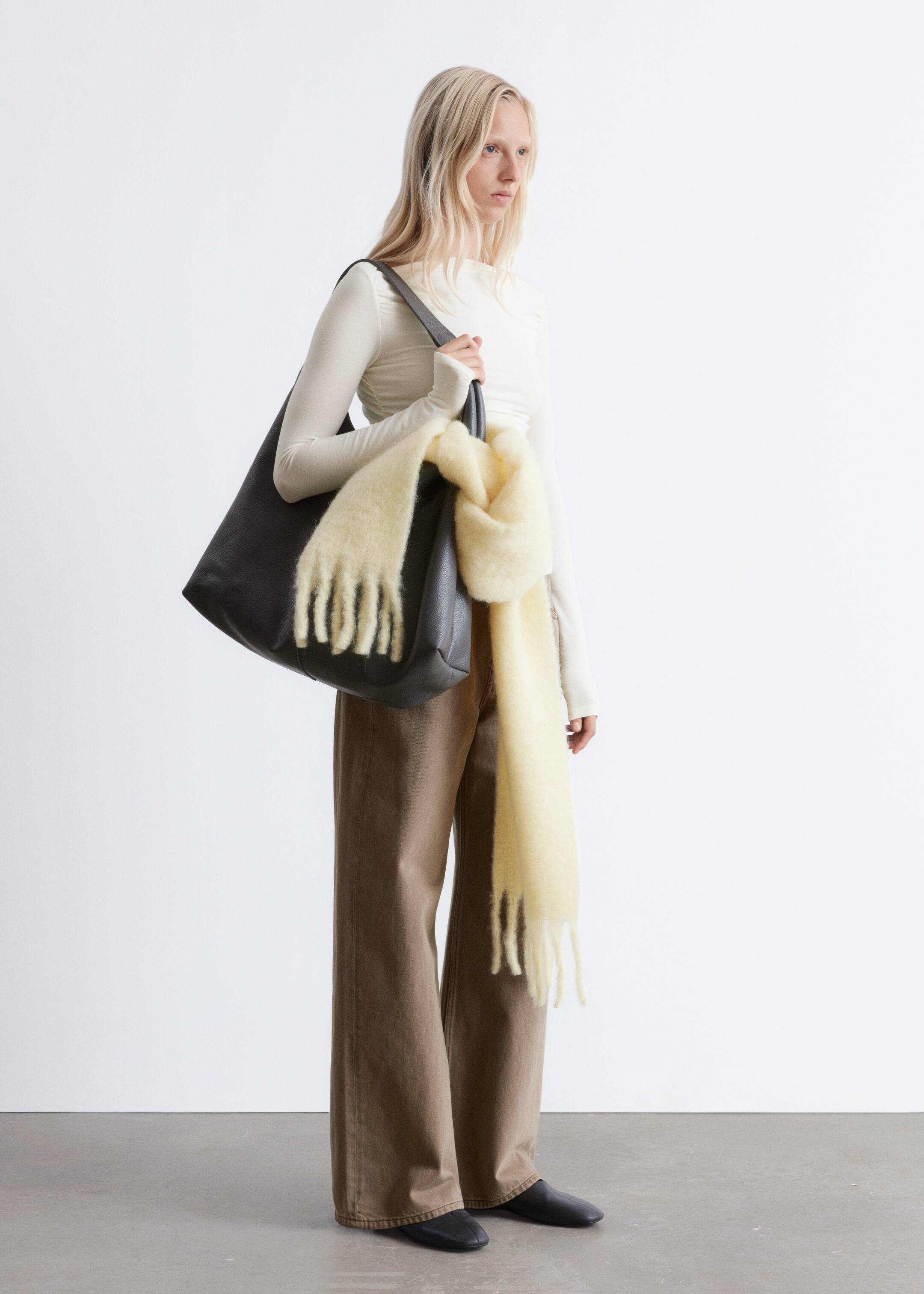 Visa större bild: En mörkgrå axelväska presenteras, hängande från en axel och vänd något framåt. Dess mjuka, ostrukturerade form är delvis dold av en ljusgul, fransad scarf draperad över framsidan.