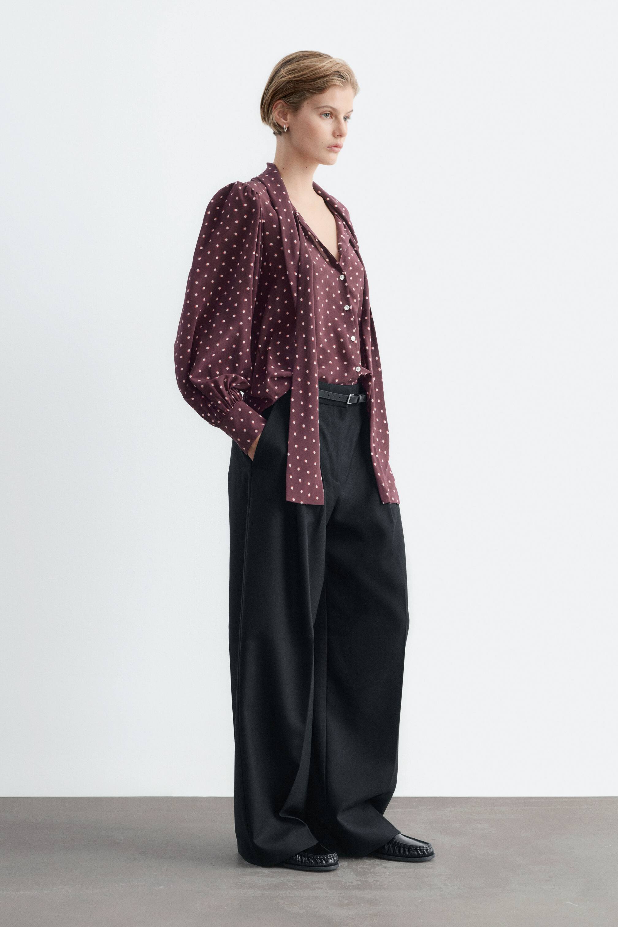 Visualizza l'immagine ingrandita: Una blusa in seta bordeaux scuro con stampa a pois bianchi viene indossata dalla modella, caratterizzata da una sciarpa annodata a doppio strato e maniche a sbuffo arricciate con polsini abbottonati. La blusa è abbinata infilata in pantaloni neri a gamba larga.