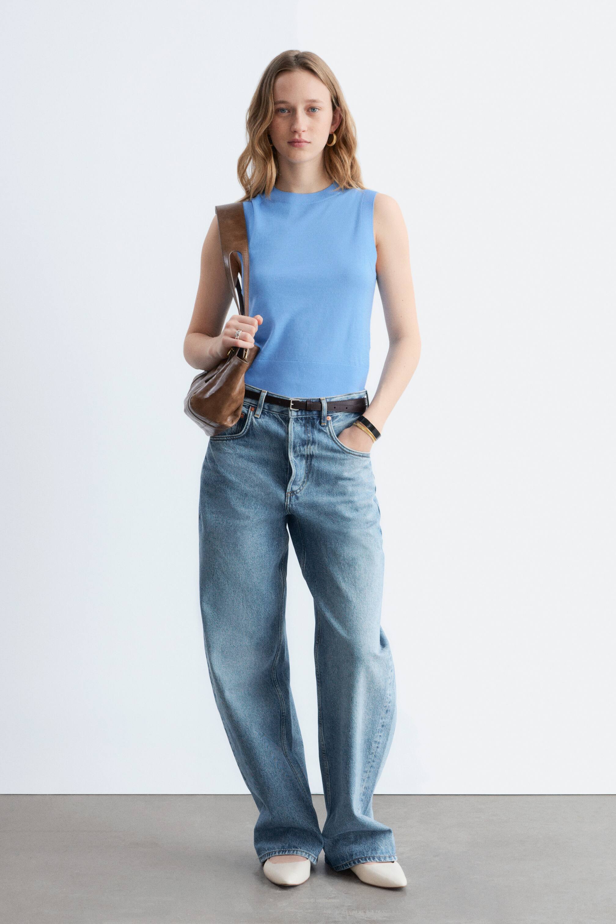 Grotere afbeelding bekijken: Een lichtblauw mouwloos topje met een ronde hals en een gebandeerde zoom wordt gedragen in een hoog getailleerde, licht gewassen blauwe wijde jeans, die voorzien is van een knoopsluiting en wordt aangesnoerd met een donkerbruine riem. Crèmekleurige puntige flats maken de outfit compleet, met een bruine leren schoudertas die over één schouder wordt gedragen.