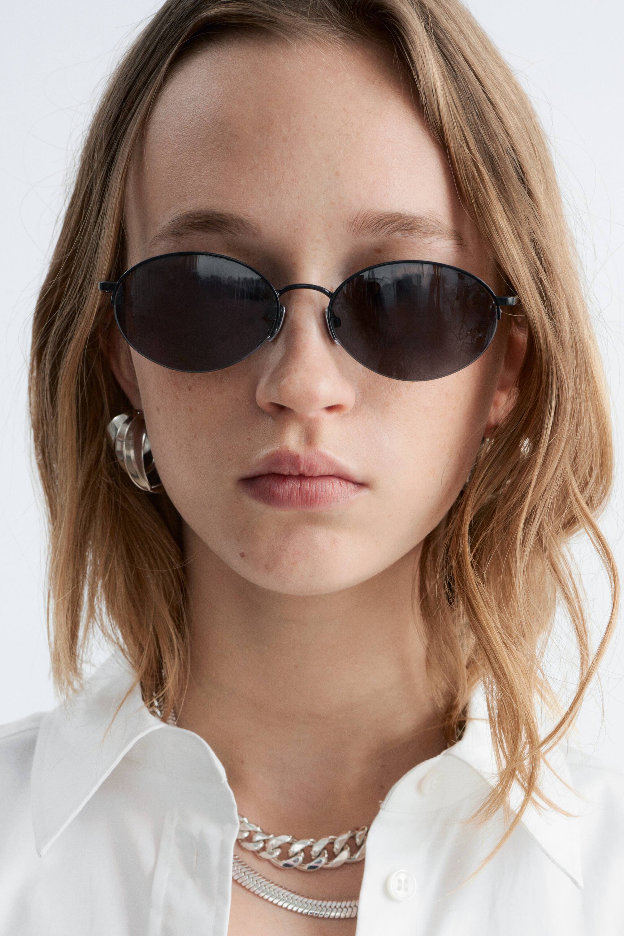 Ver imagen más grande: Las gafas de sol presentan lentes ovaladas con tintado oscuro y finas monturas de metal negro, llevadas en el rostro de una persona y orientadas hacia adelante.