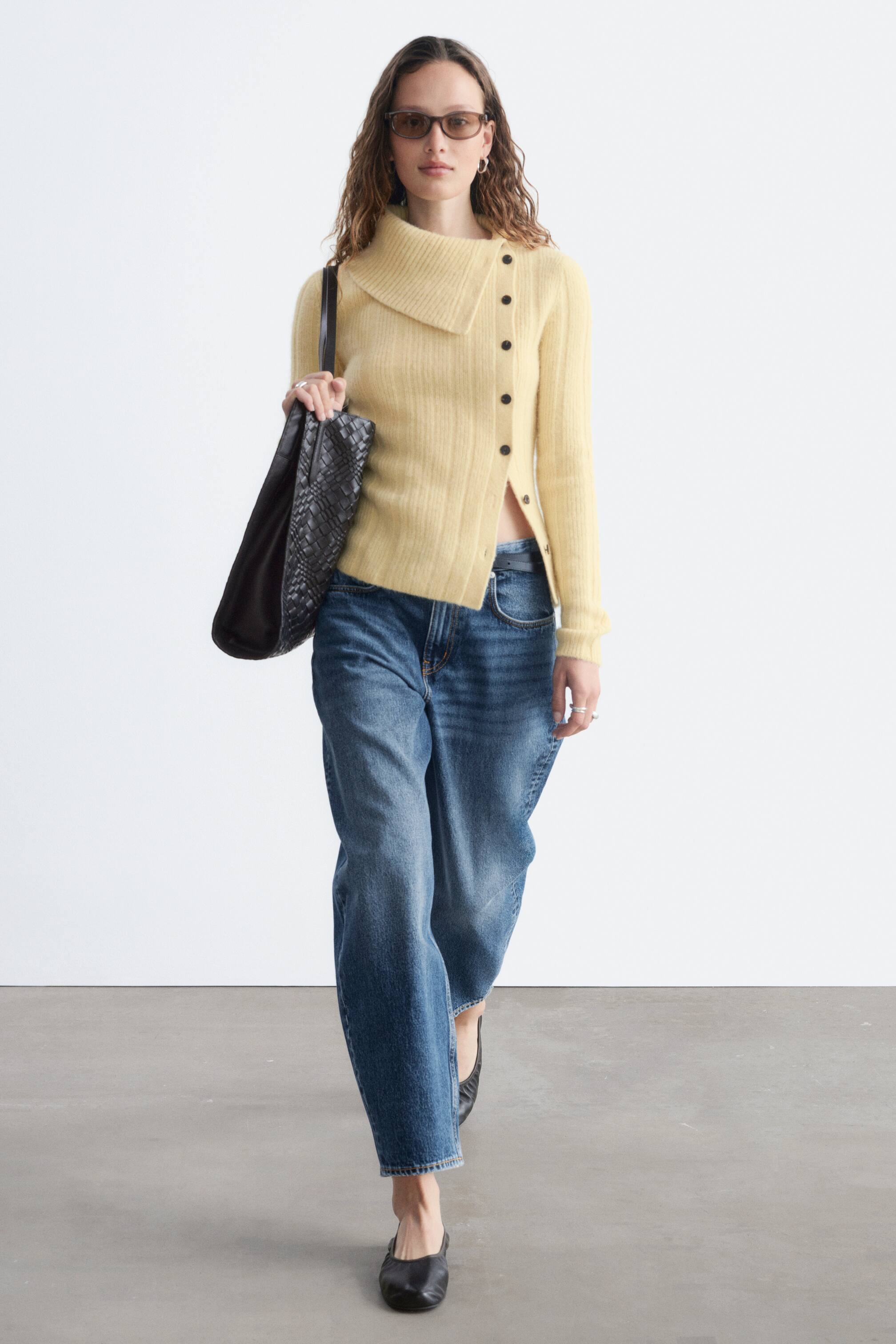 View larger image: A light yellow fuzzy knit cardigan features an asymmetrical design, showcasing a high, cowl-like collar on the left side and a diagonal button fastening with dark buttons. It has long sleeves and a cropped, asymmetrical hem.