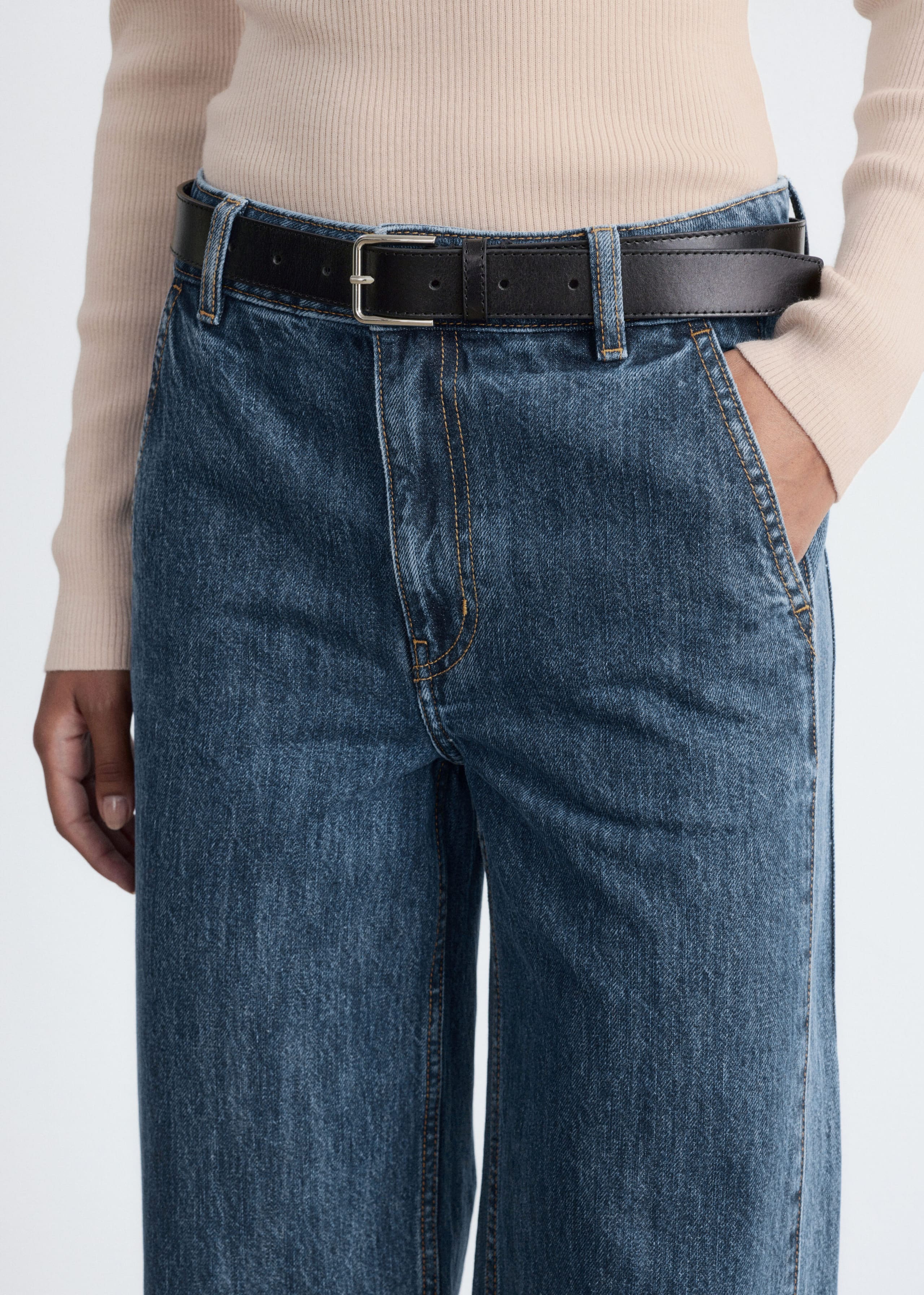 The model is wearing high-waisted, wide-leg blue denim jeans, with one hand resting in a slanted side pocket. The jeans are styled with a black leather belt and a tucked-in beige ribbed top.