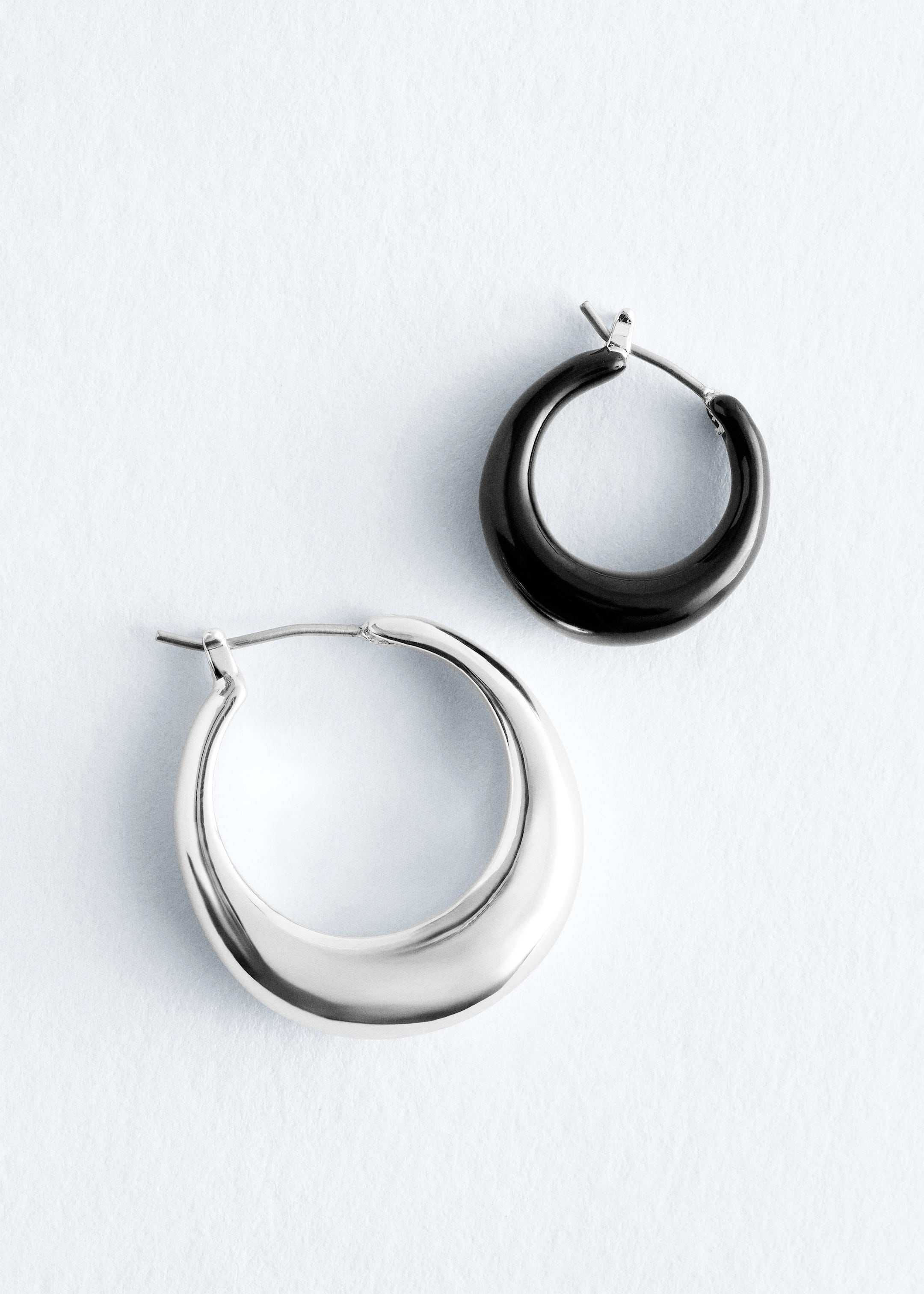 Größeres Bild anzeigen: Ein Set aus zwei Creolen liegt flach, bestehend aus einem größeren Silberohrring und einem kleineren schwarzen Ohrring. Beide Ohrringe verbreitern sich nach unten und sind mit einem Scharnier-Verschluss gestaltet.